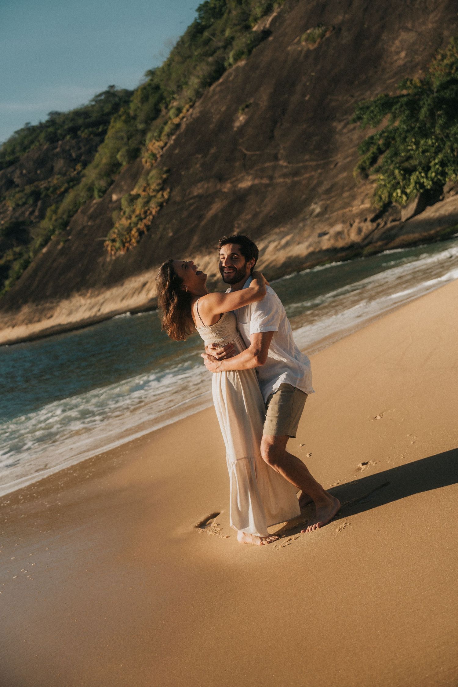 A couple shares a romantic moment on a scenic beach with rocky cliffs and ocean waves in the background.