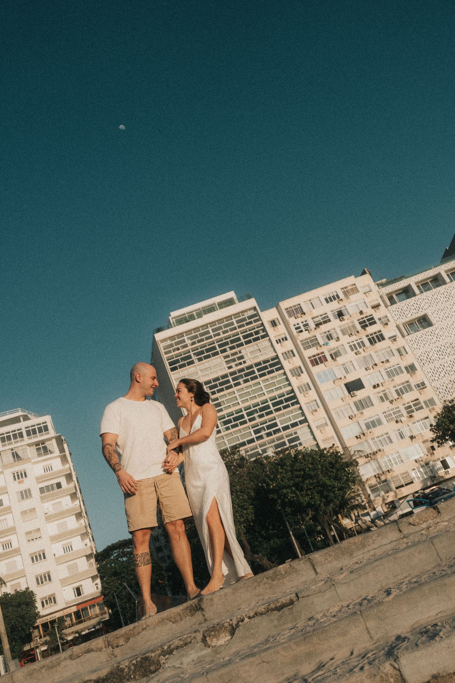 A couple stands together on a beach with high-rise apartment buildings in the background on a sunny day.