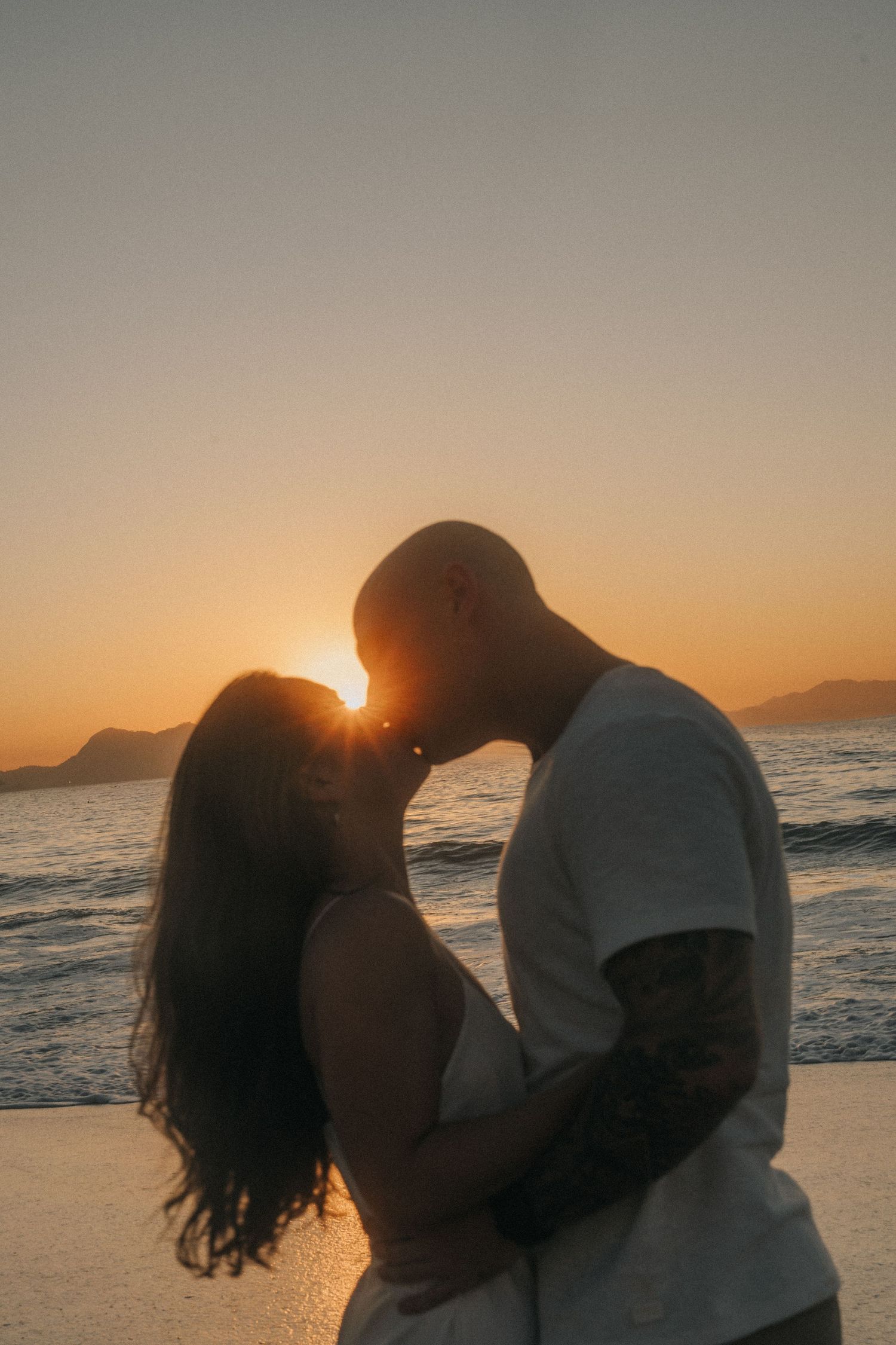 Silhouetted couple sharing intimate moments on beach against orange sunset sky as sun sets over ocean horizon.
