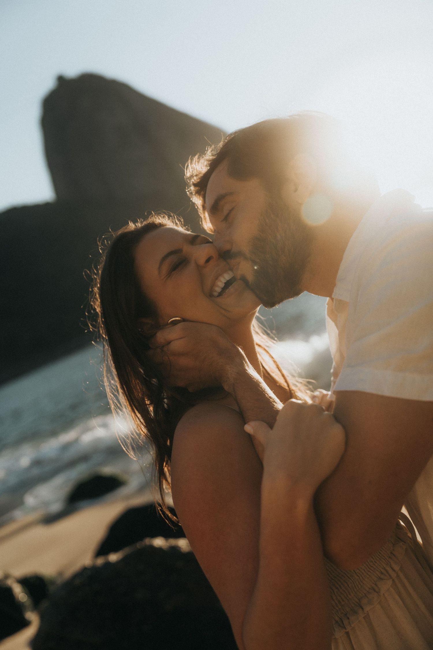 Romantic couple share intimate moment at sunset on a beach with golden light and cliffs in background.