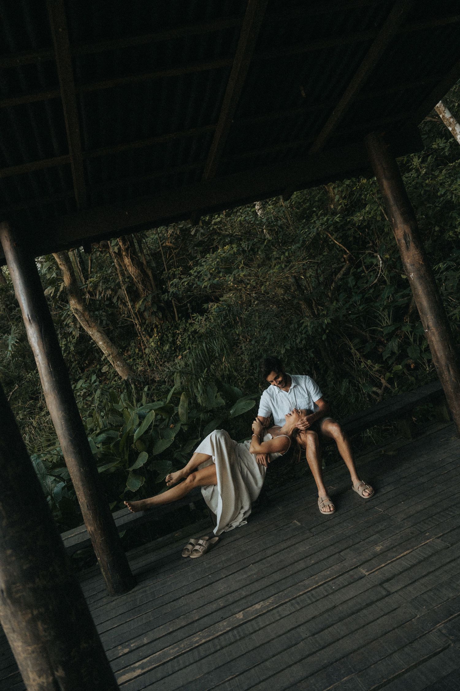 A couple embraces intimately on a wooden deck surrounded by lush trees and greenery in a moody setting.