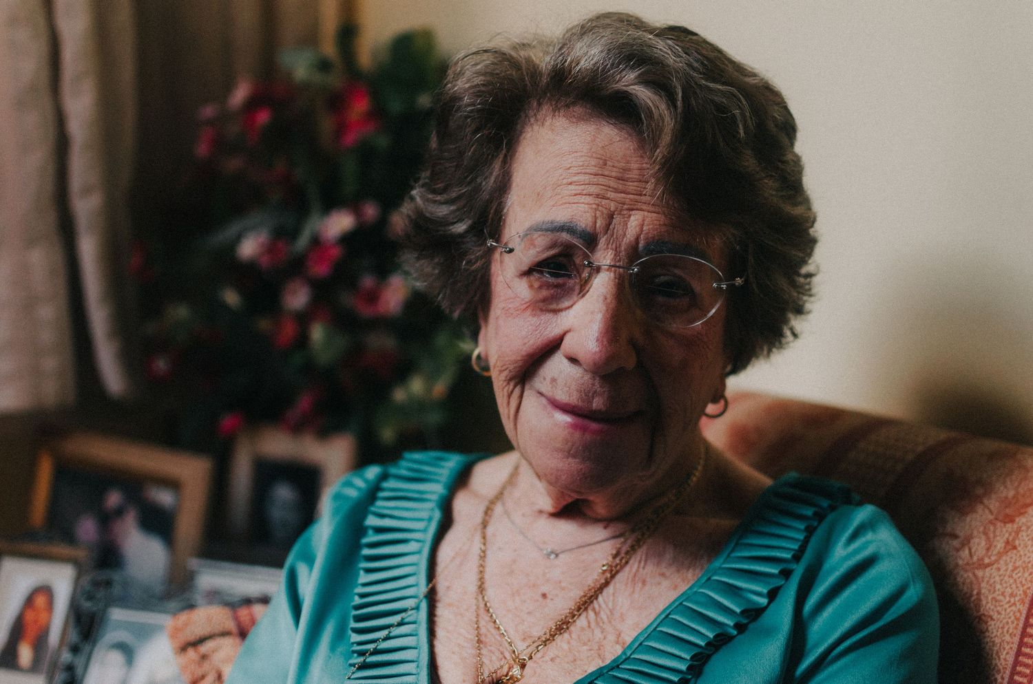 Elderly woman in teal cardigan sits comfortably at home with a warm smile and curly hair.