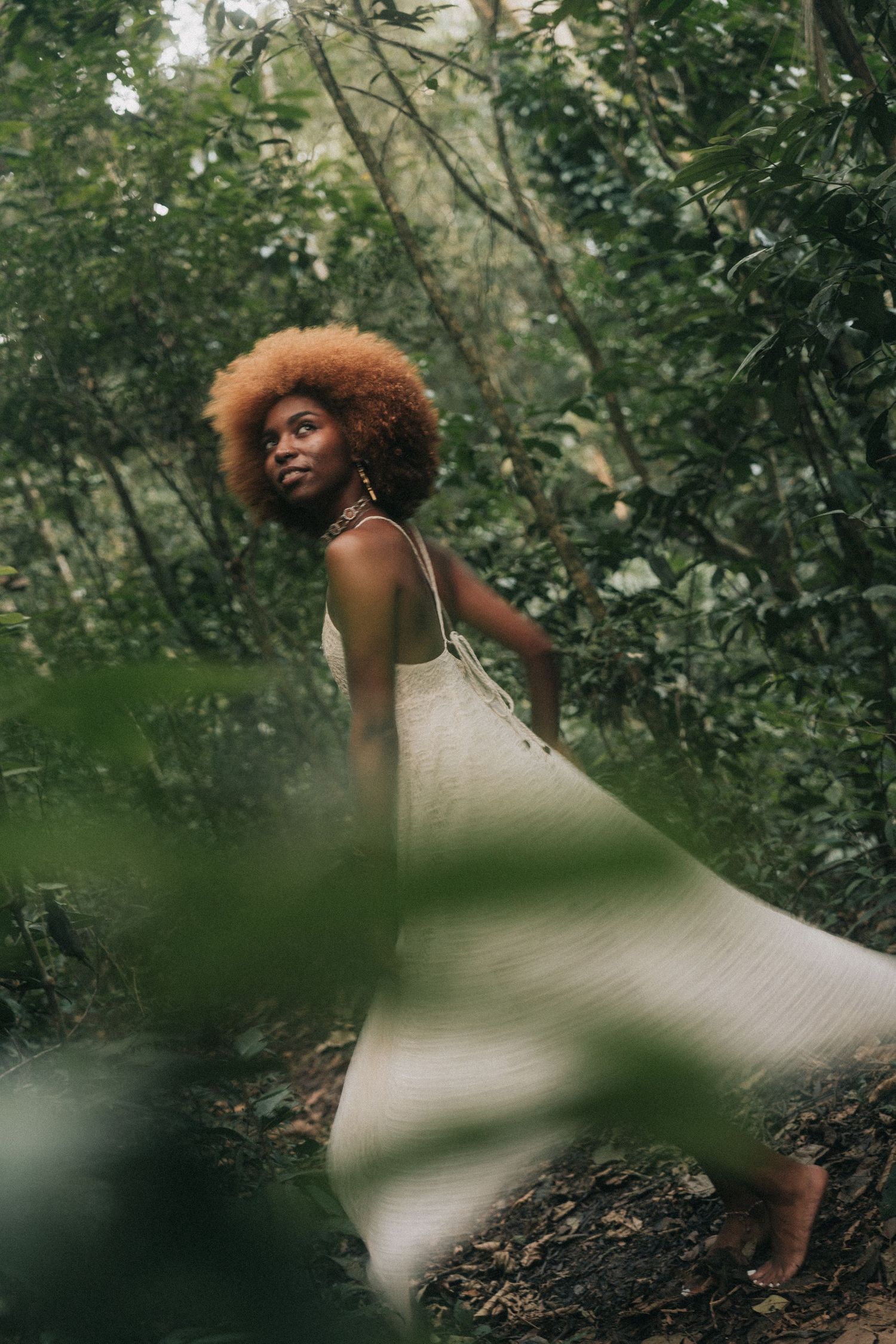A person in a flowing white dress poses dramatically among lush green foliage in an artistic outdoor photo series.