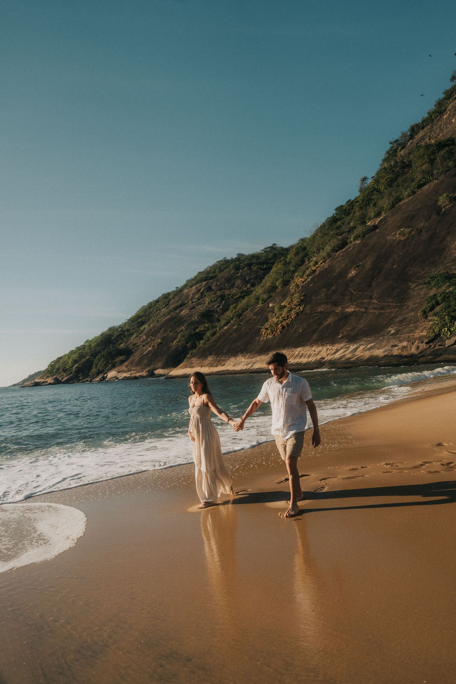 Two people walk hand in hand along a scenic golden sandy beach with cliffs and ocean waves in the background.