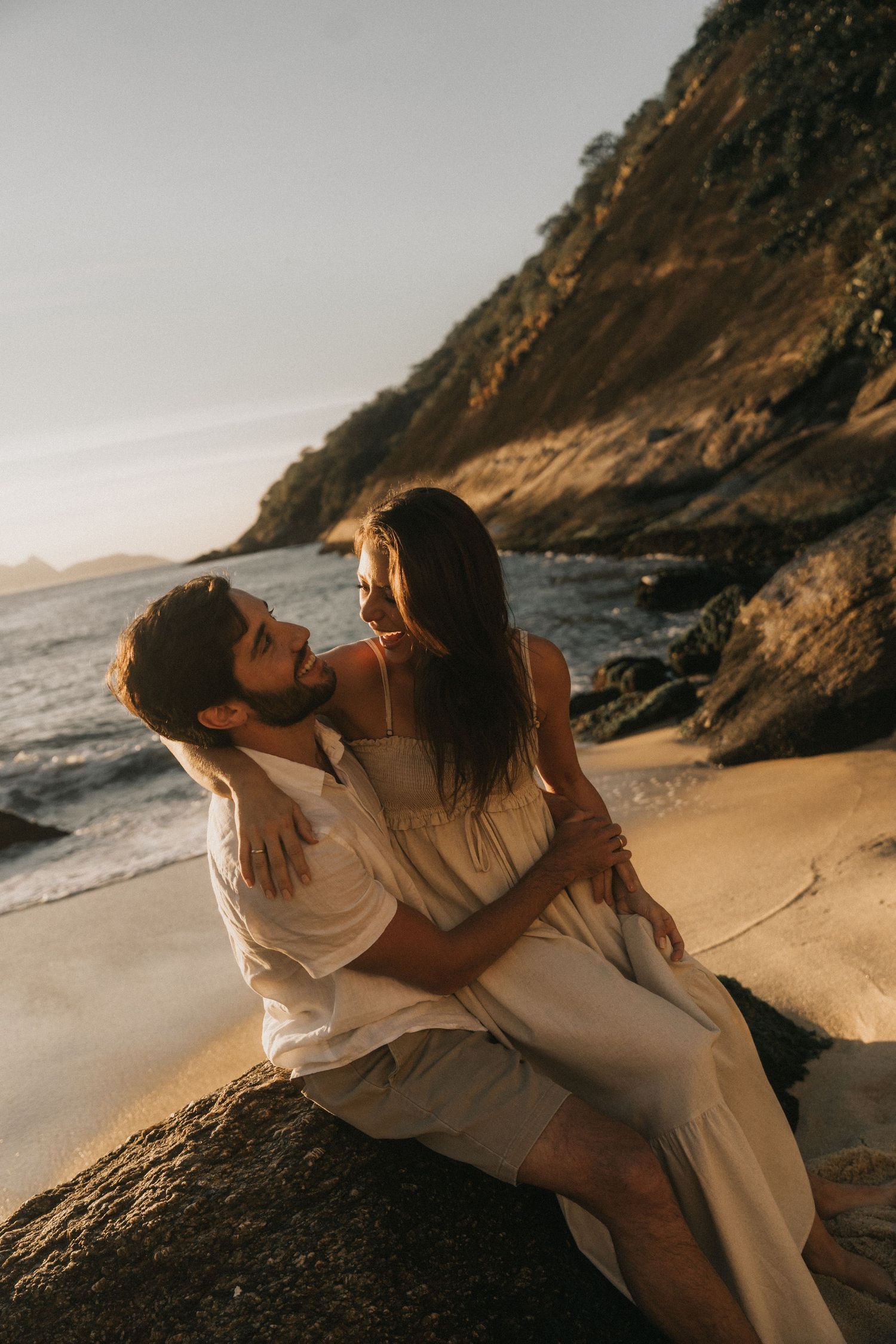 Two people share an intimate embrace on a sandy beach with dramatic cliffs and ocean waves at sunset.