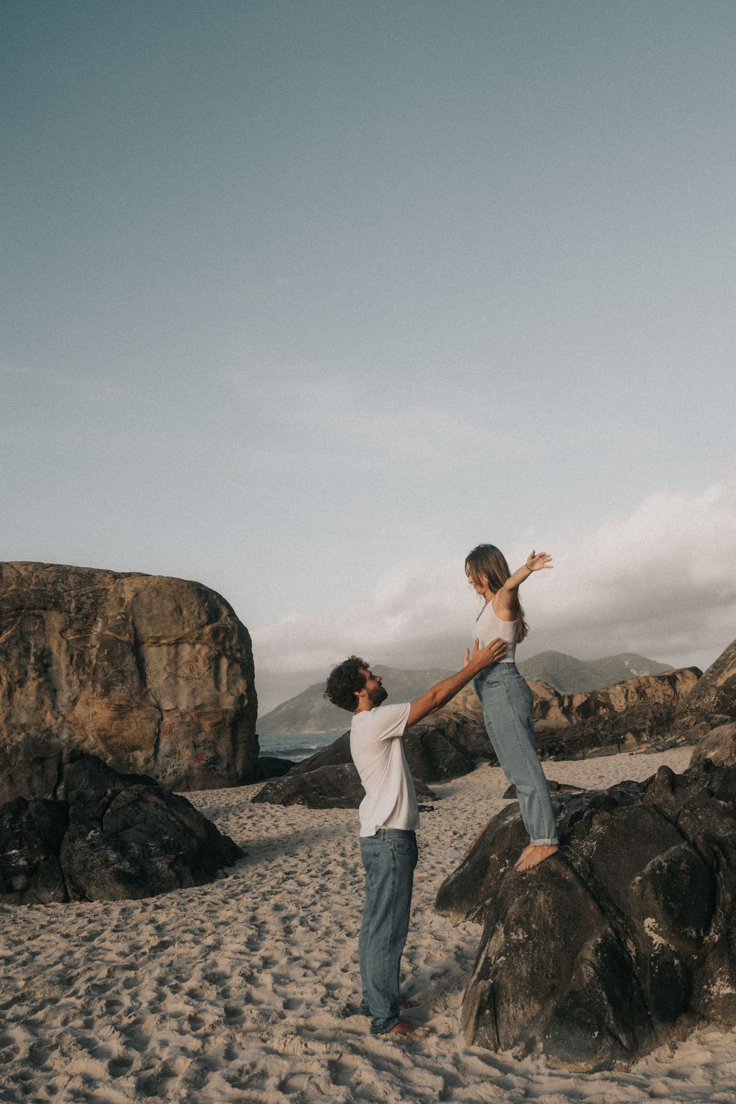 Two people playfully explore a rocky coastal beach against dramatic cliffs and cloudy skies at sunset.