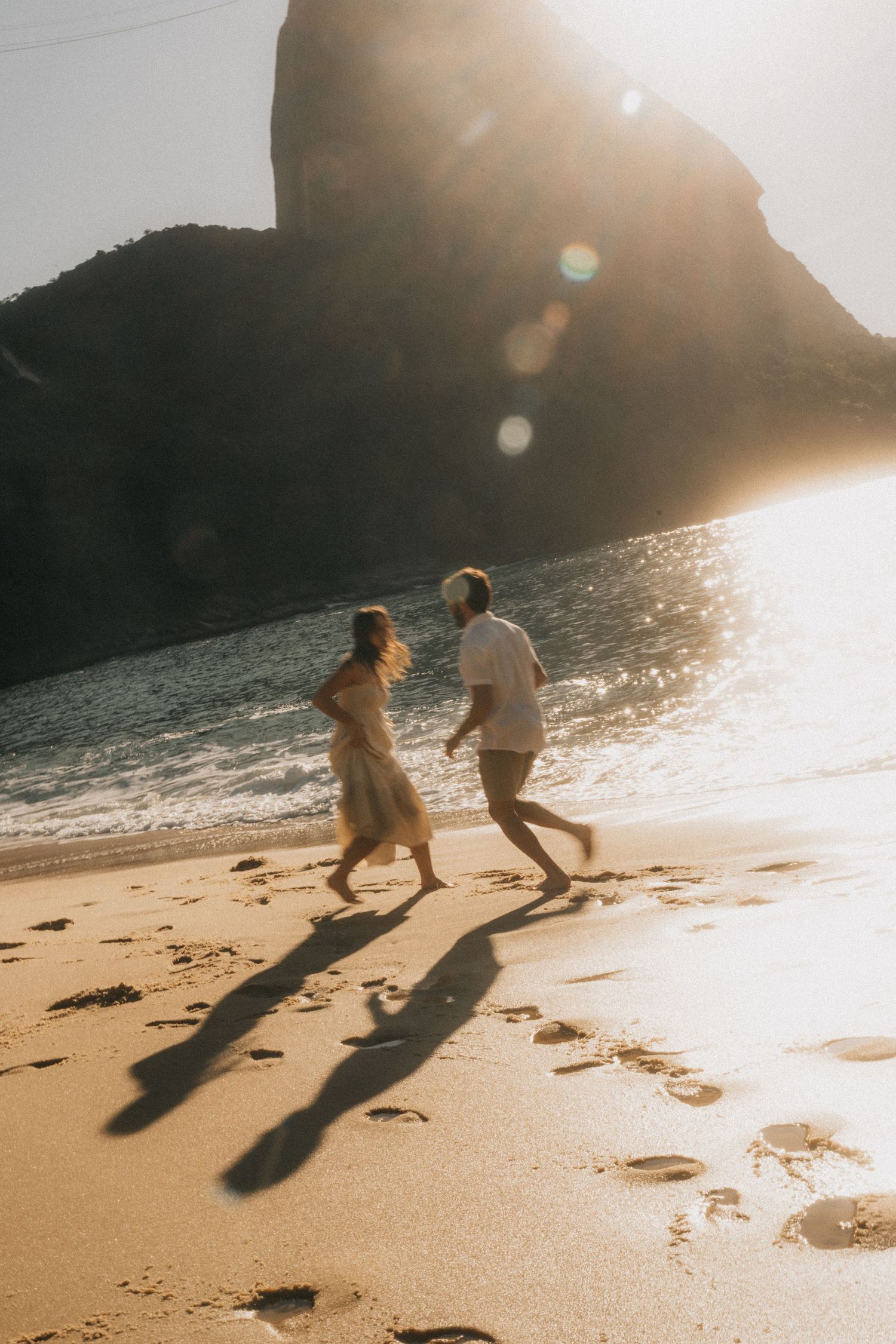 Couple running together on a sandy beach at sunset with dramatic shadows cast on the sand as sun peeks over cliffs.