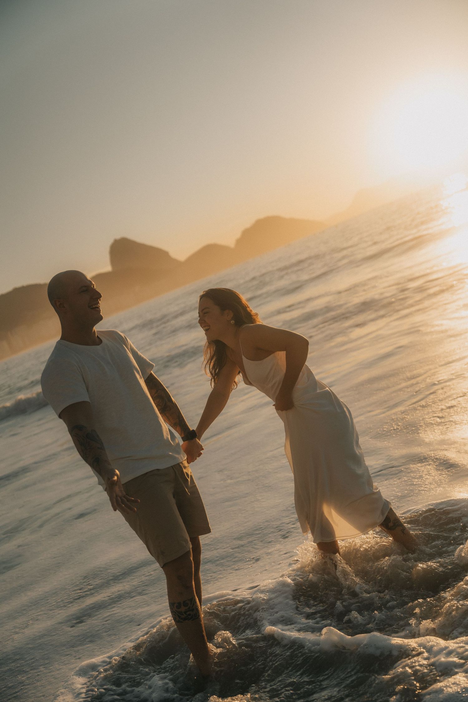A silhouetted couple holds hands while standing in ocean waves during a romantic sunset at the beach.