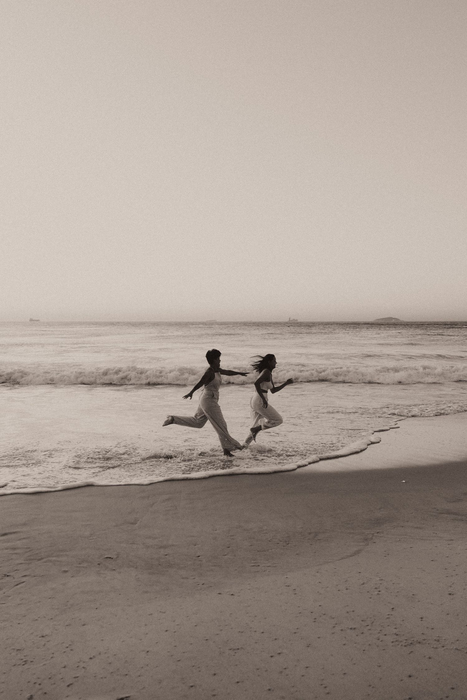 Two silhouetted figures playfully run along a misty beach as ocean waves roll in during sunset, captured in black and white.