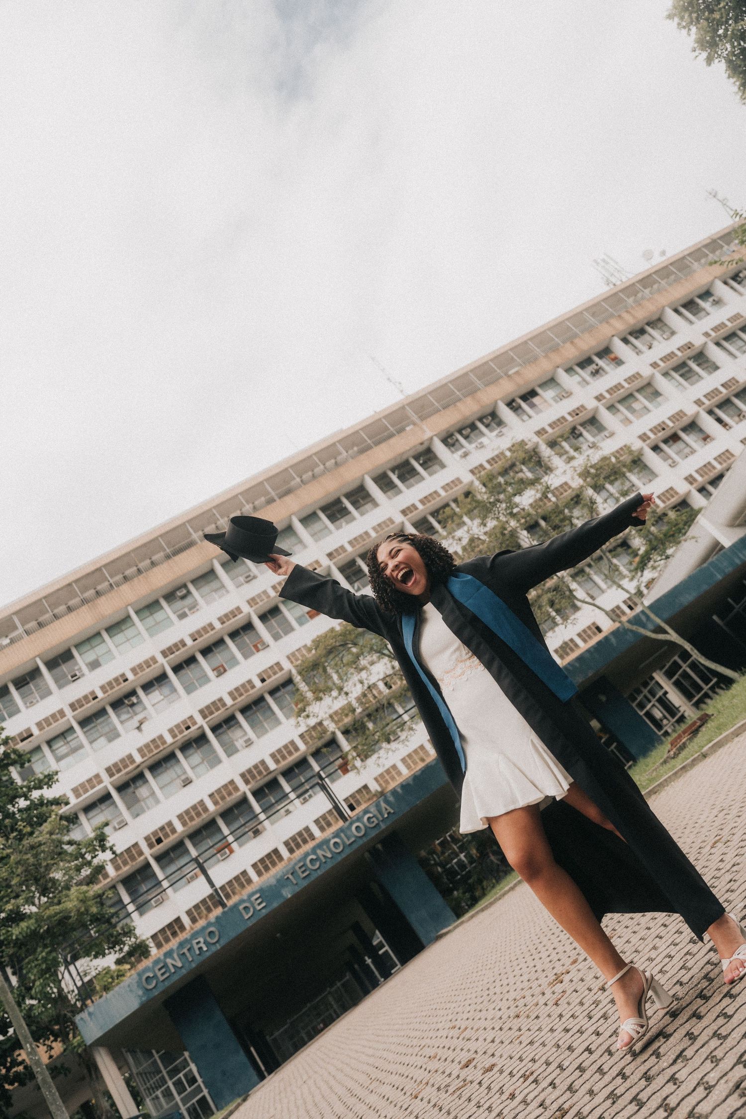 A person in graduation regalia celebrates outdoors in front of a large academic building with their arms raised joyfully.