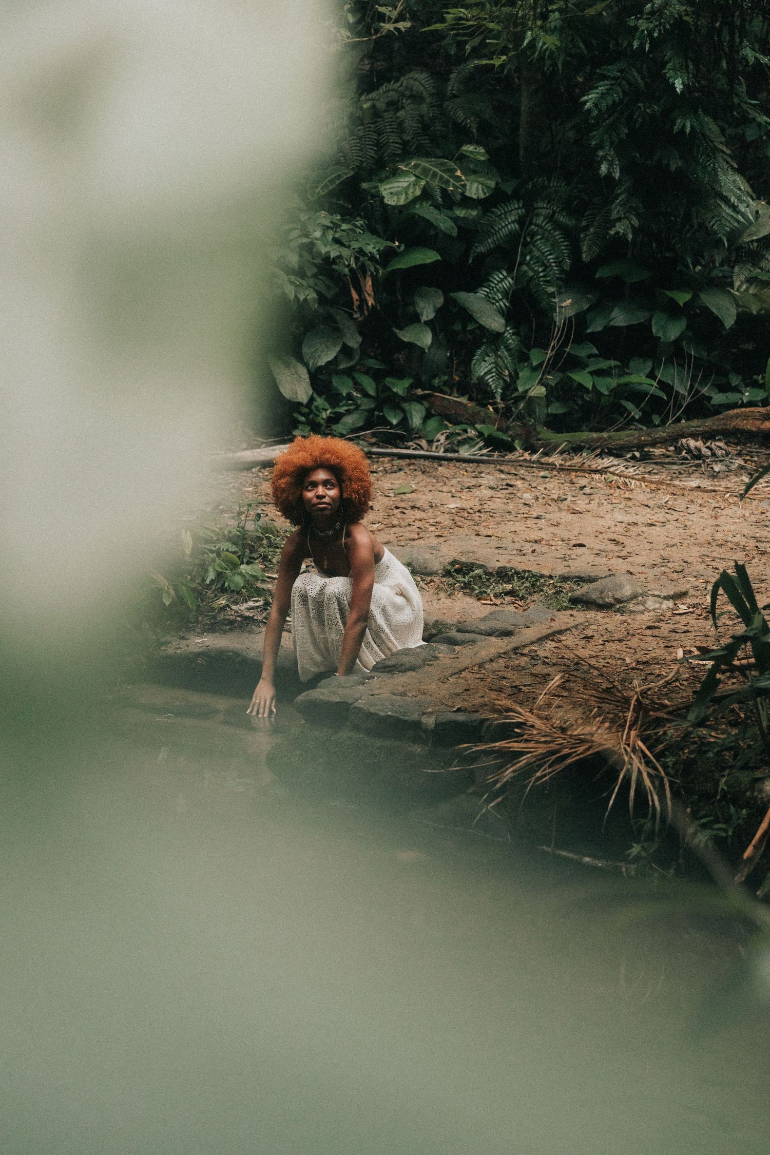 Person in traditional dress carrying a large bundle of straw or hay through a lush green garden setting.