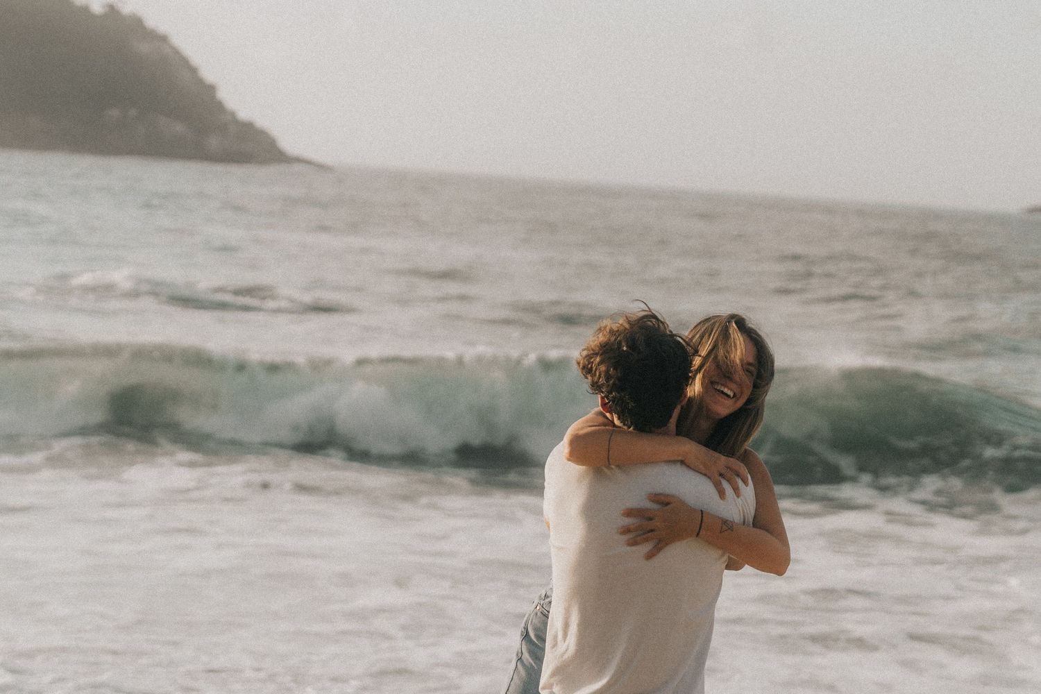 A couple embraces while standing on a rocky beach with crashing ocean waves in the background.