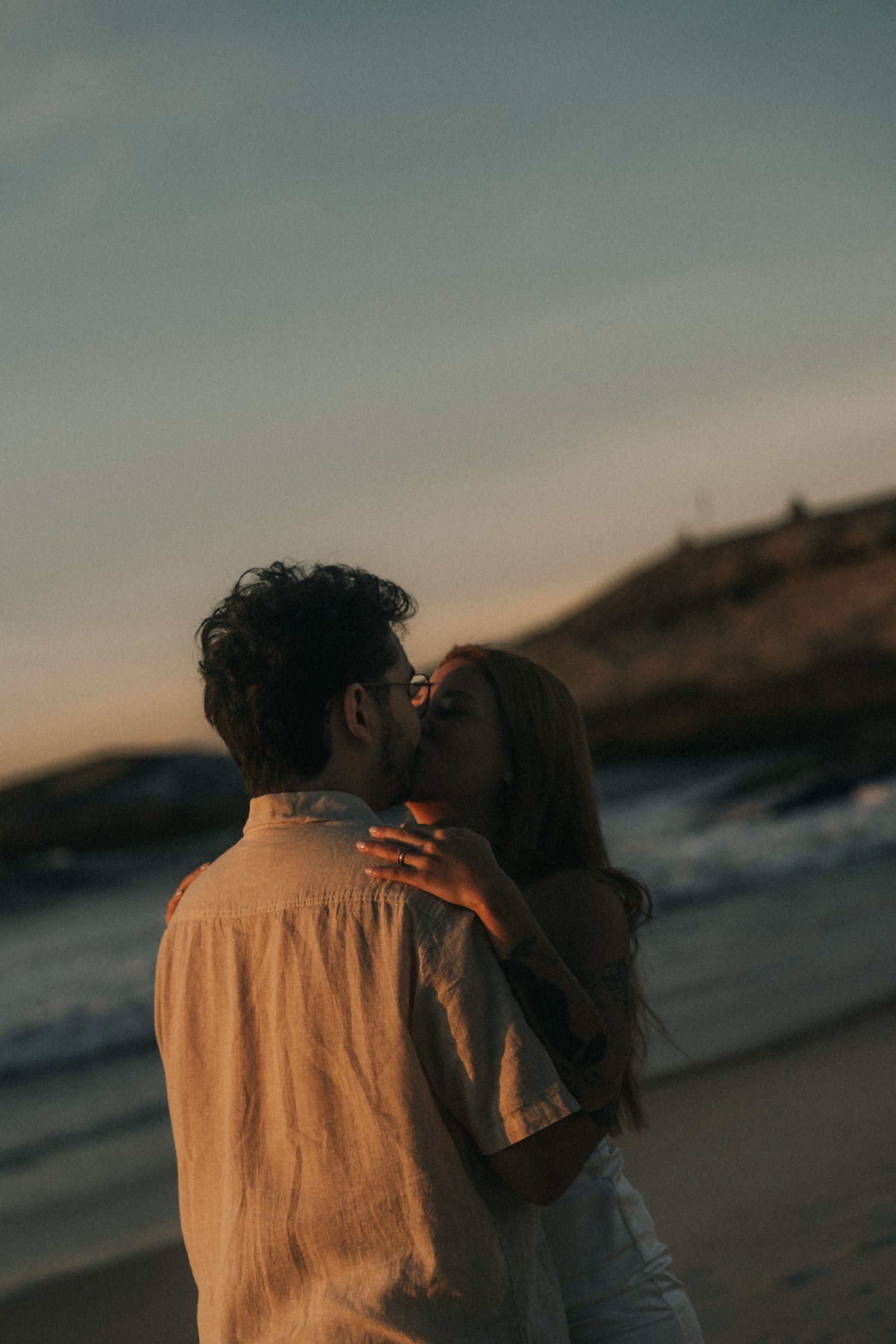 A couple shares an intimate kiss on the beach at sunset with waves in the background.