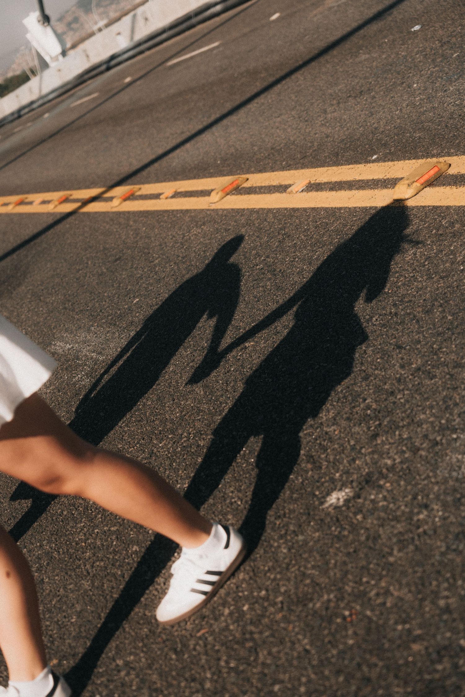 Long shadows of two people cast on asphalt next to yellow caution tape, with a person's foot wearing white sneakers visible.
