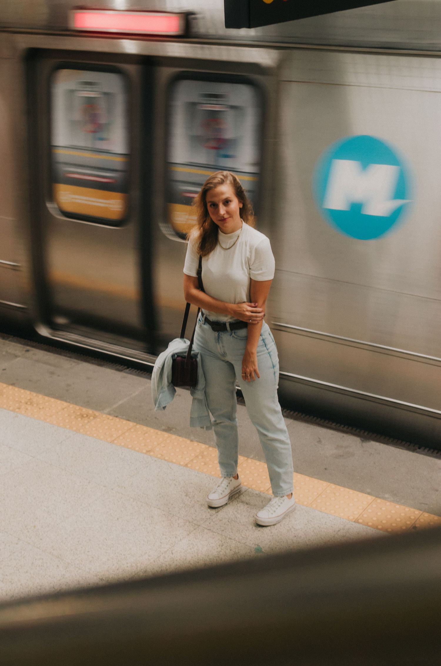 Person in white sneakers and gray pants stands on subway platform near train with motion blur effect.