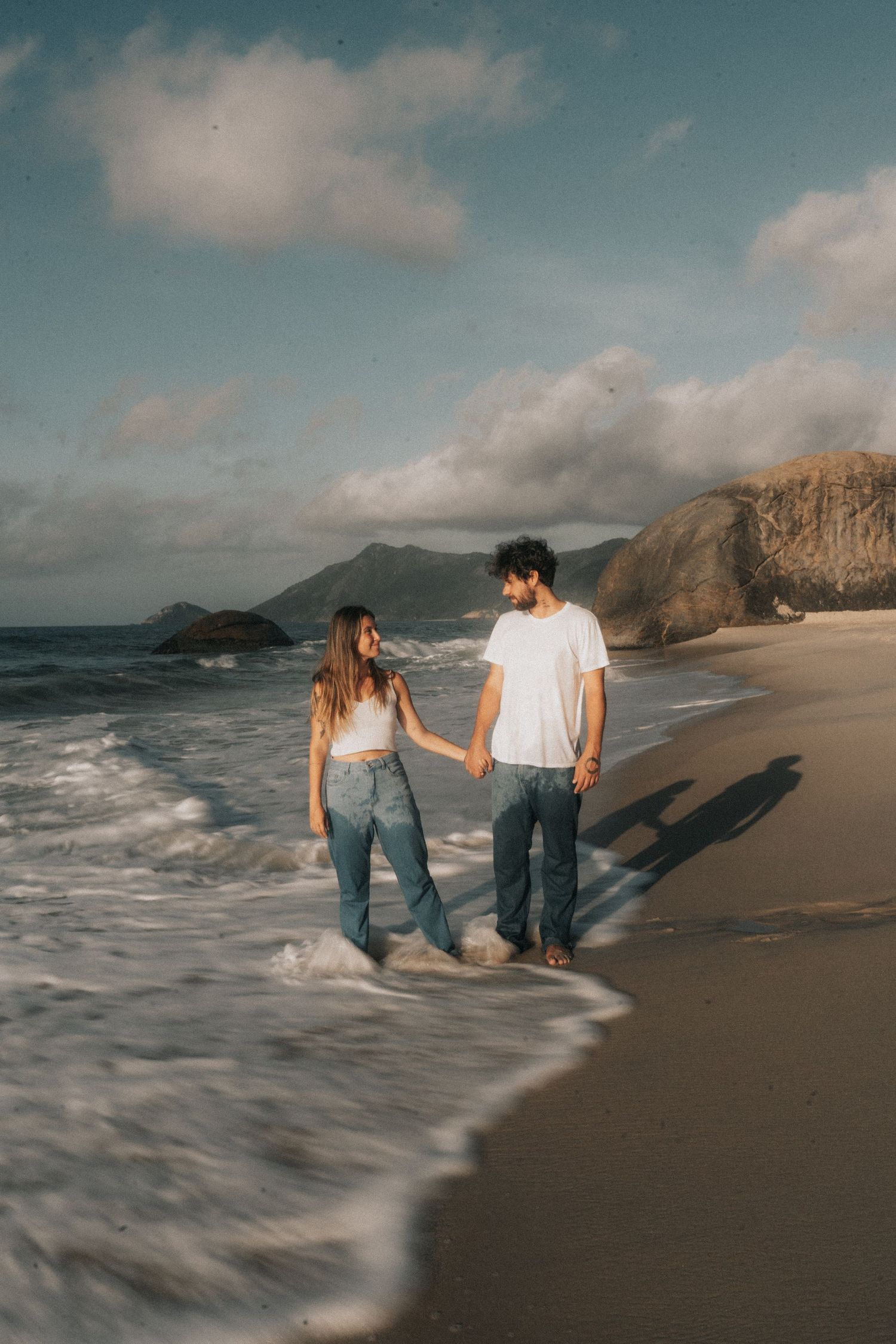 A couple walks along a scenic beach with cliffs and waves during golden hour on the California coast.