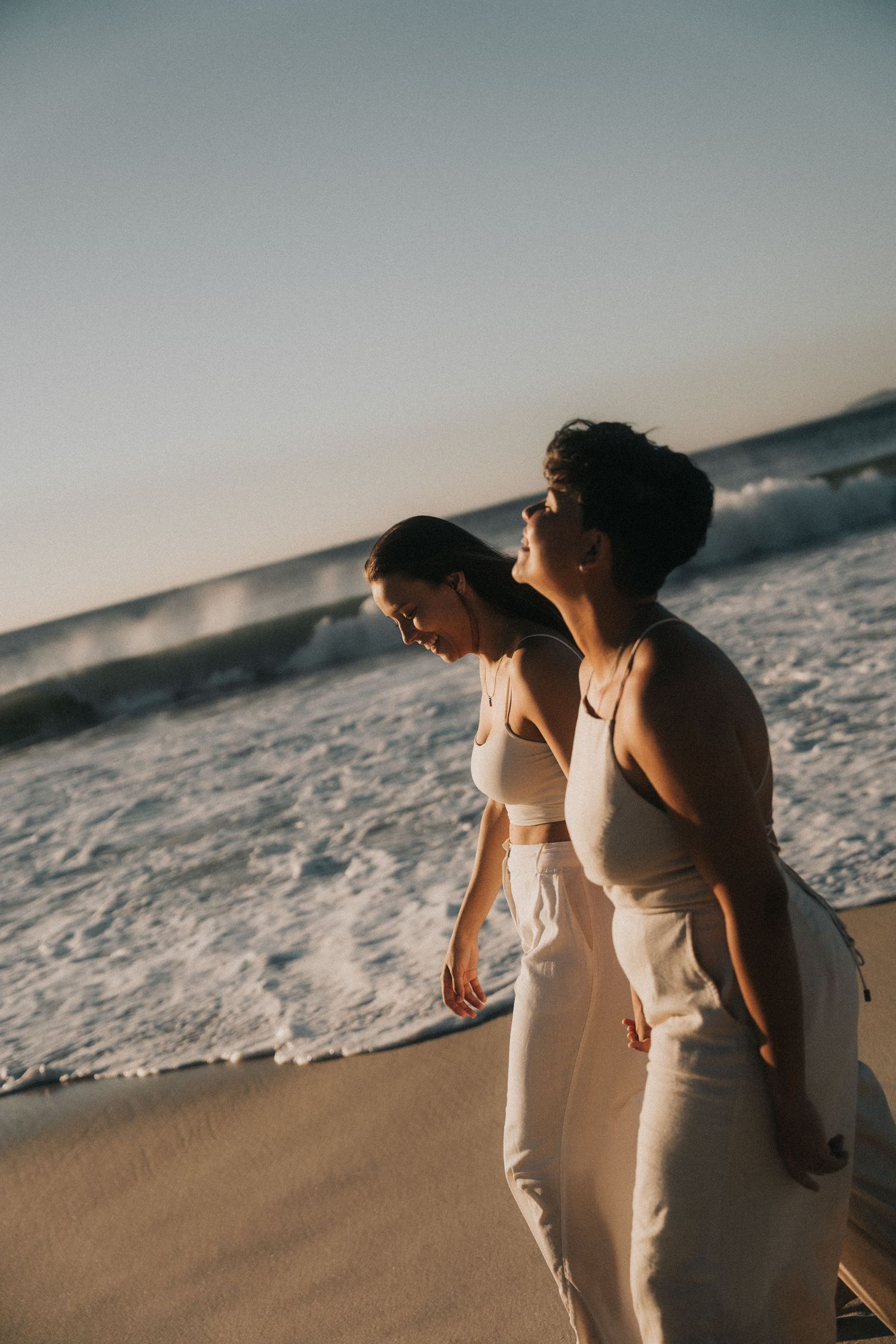 Two figures walk together on a sandy beach at sunset casting long shadows across the shoreline.