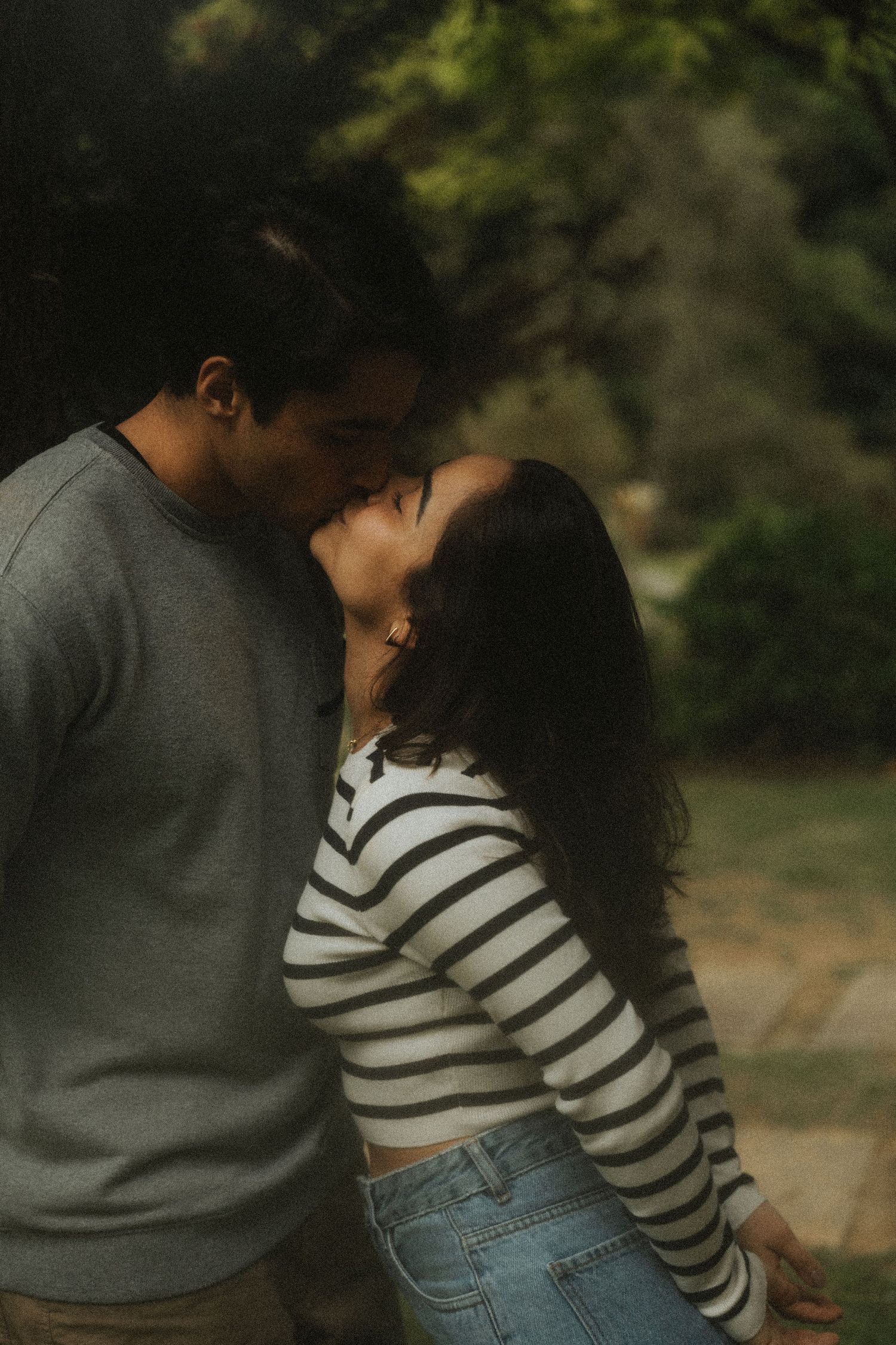 A romantic couple in casual clothing share an intimate moment outdoors with natural lighting and greenery in background.