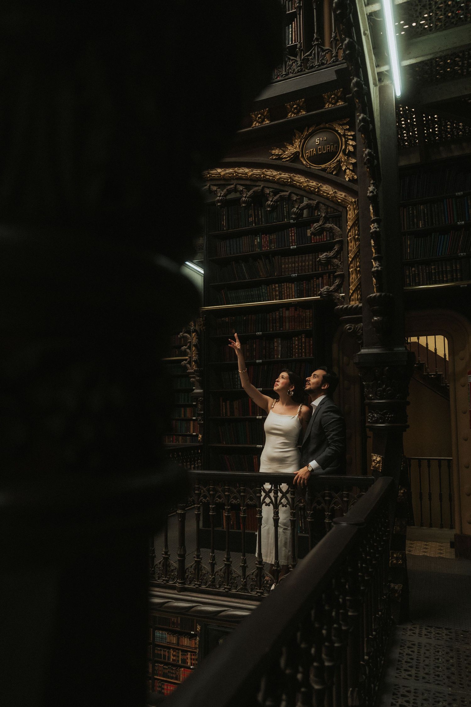 A couple poses dramatically on ornate vintage library stairs with carved wooden details and dramatic lighting.