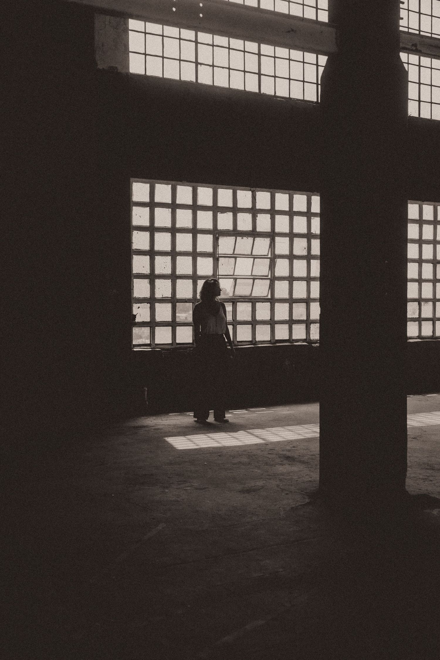 A dark, minimalist interior space with industrial windows casting dramatic shadows on an empty floor.