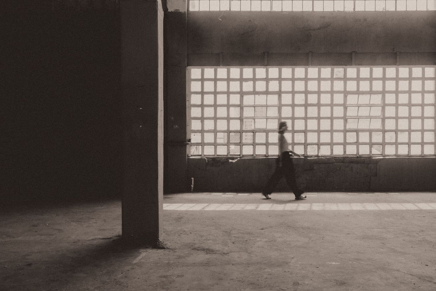 A silhouetted figure walks past a geometric window pattern in a minimalist architectural space.