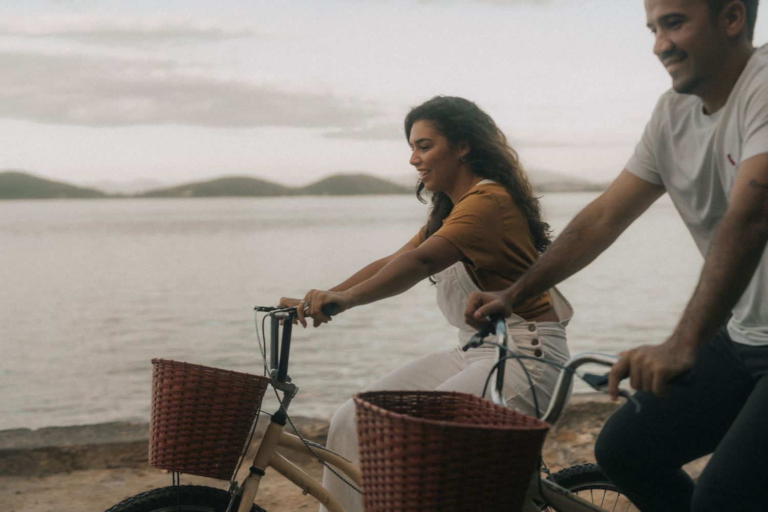 A couple rides bicycles together along a shoreline path at sunset creating a romantic outdoor scene.