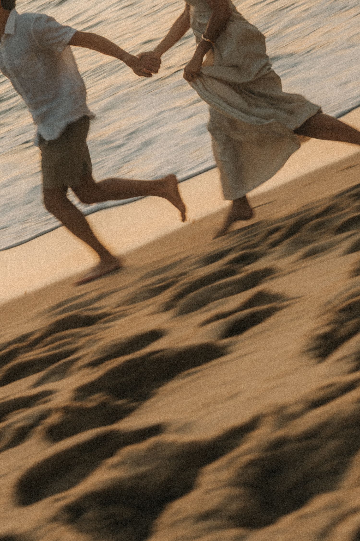 Two figures in flowing white clothes holding hands and running together on rippling sand dunes at sunset.