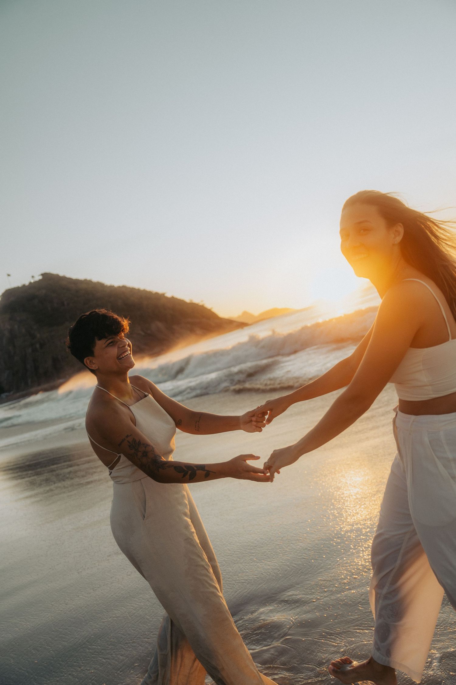 Two figures in white outfits dance and twirl together on a beach at sunset with waves crashing behind them.