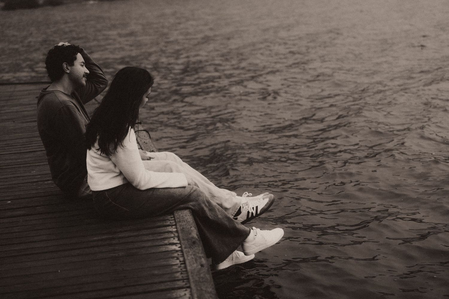 A romantic couple sits together on a wooden dock by the water in black and white photography.