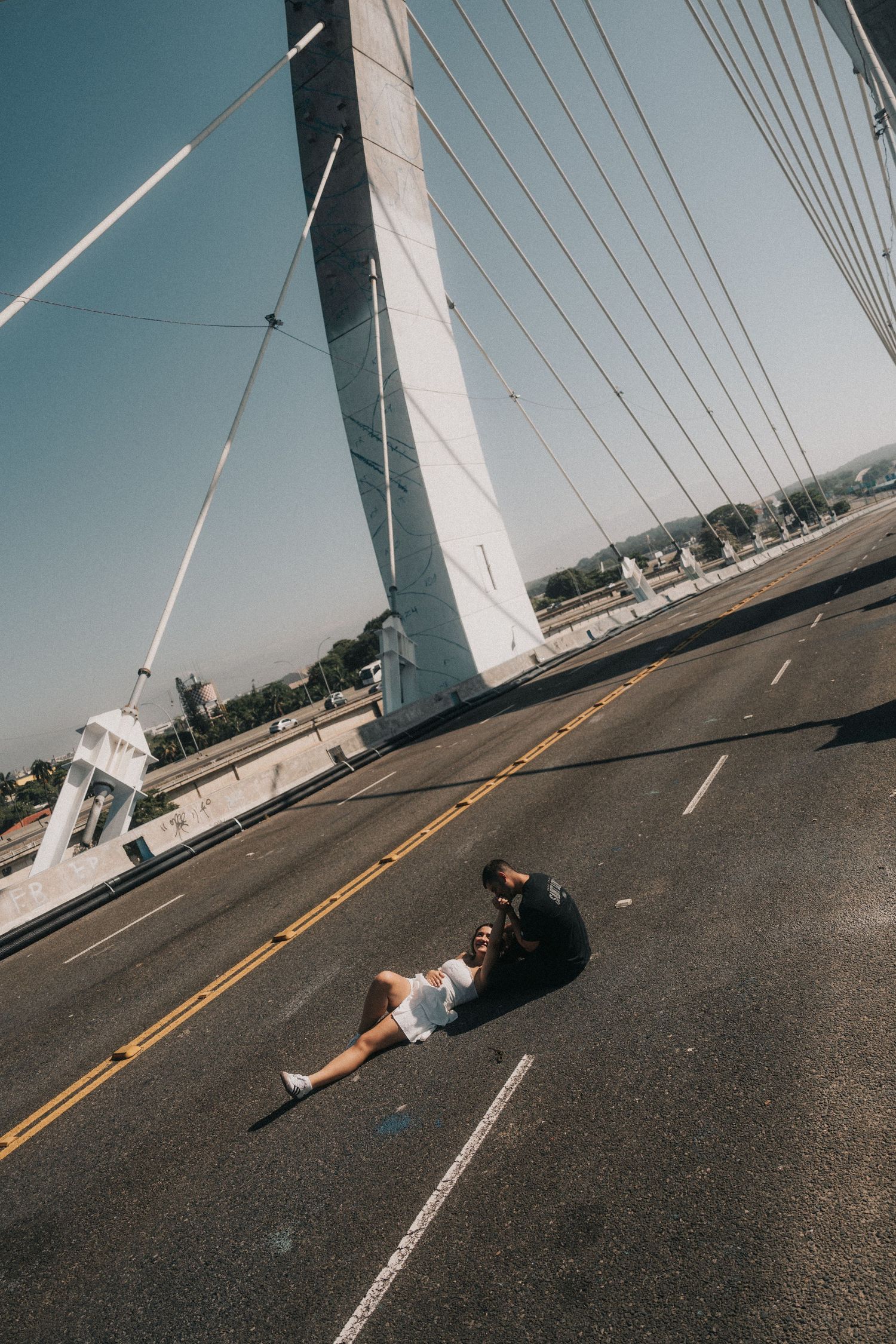 Person lying on asphalt road with cable bridge cables visible against blue sky and white support tower.