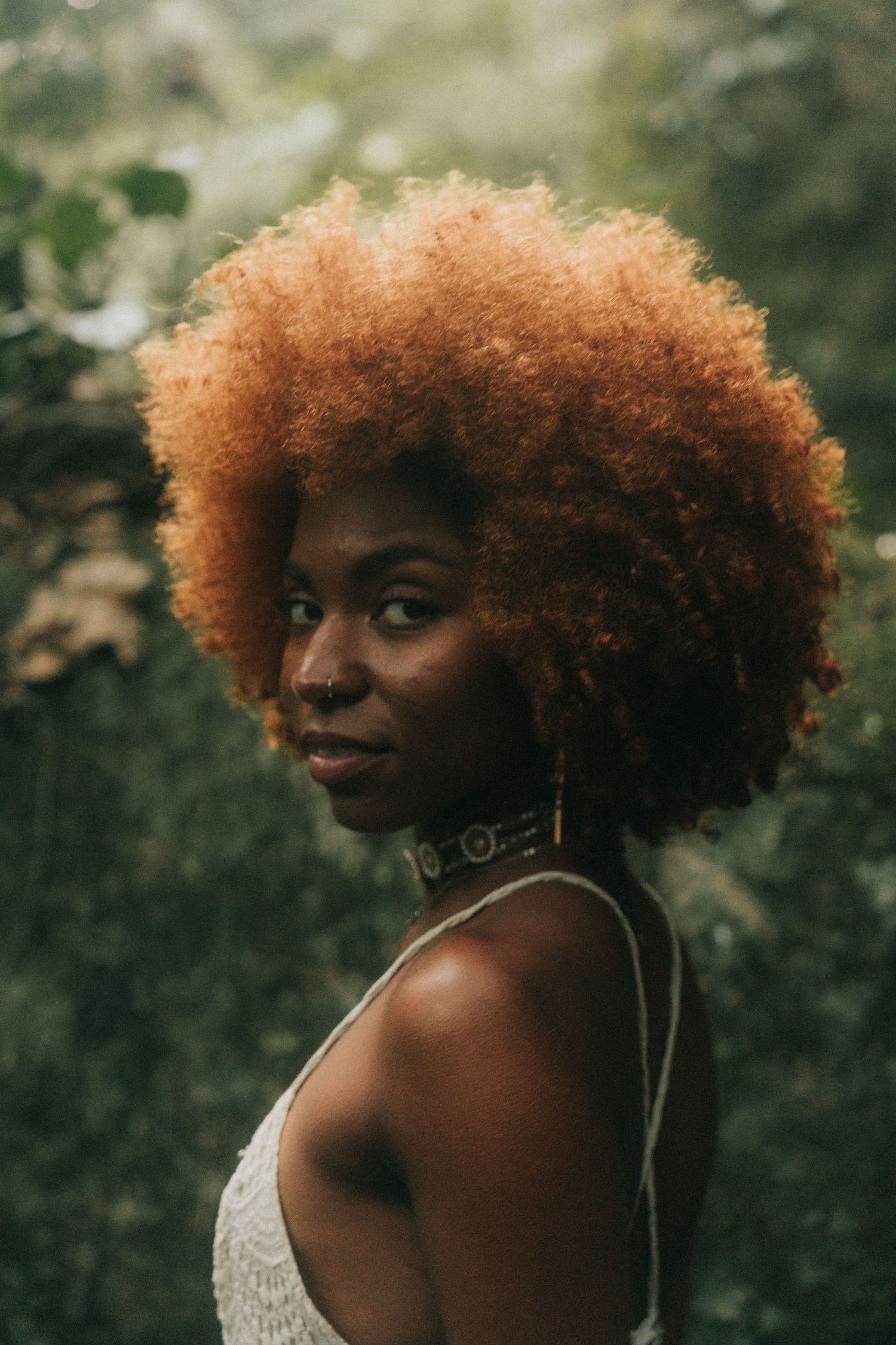 Portrait against green foliage showing vibrant copper-colored natural curls and white lace top in warm natural lighting.