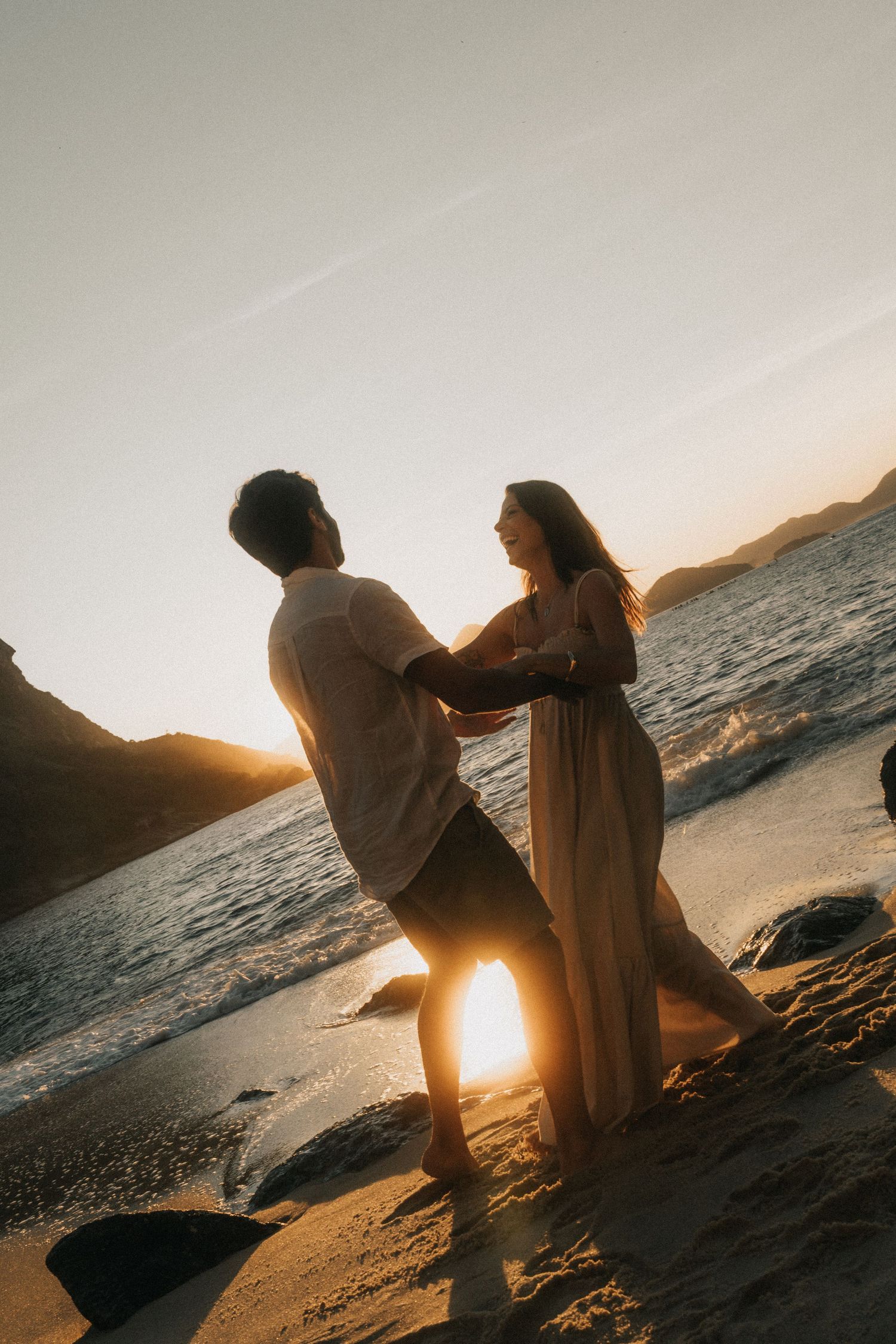 Two silhouetted figures share an intimate moment on a beach at sunset with rays of sunlight streaming through between them.