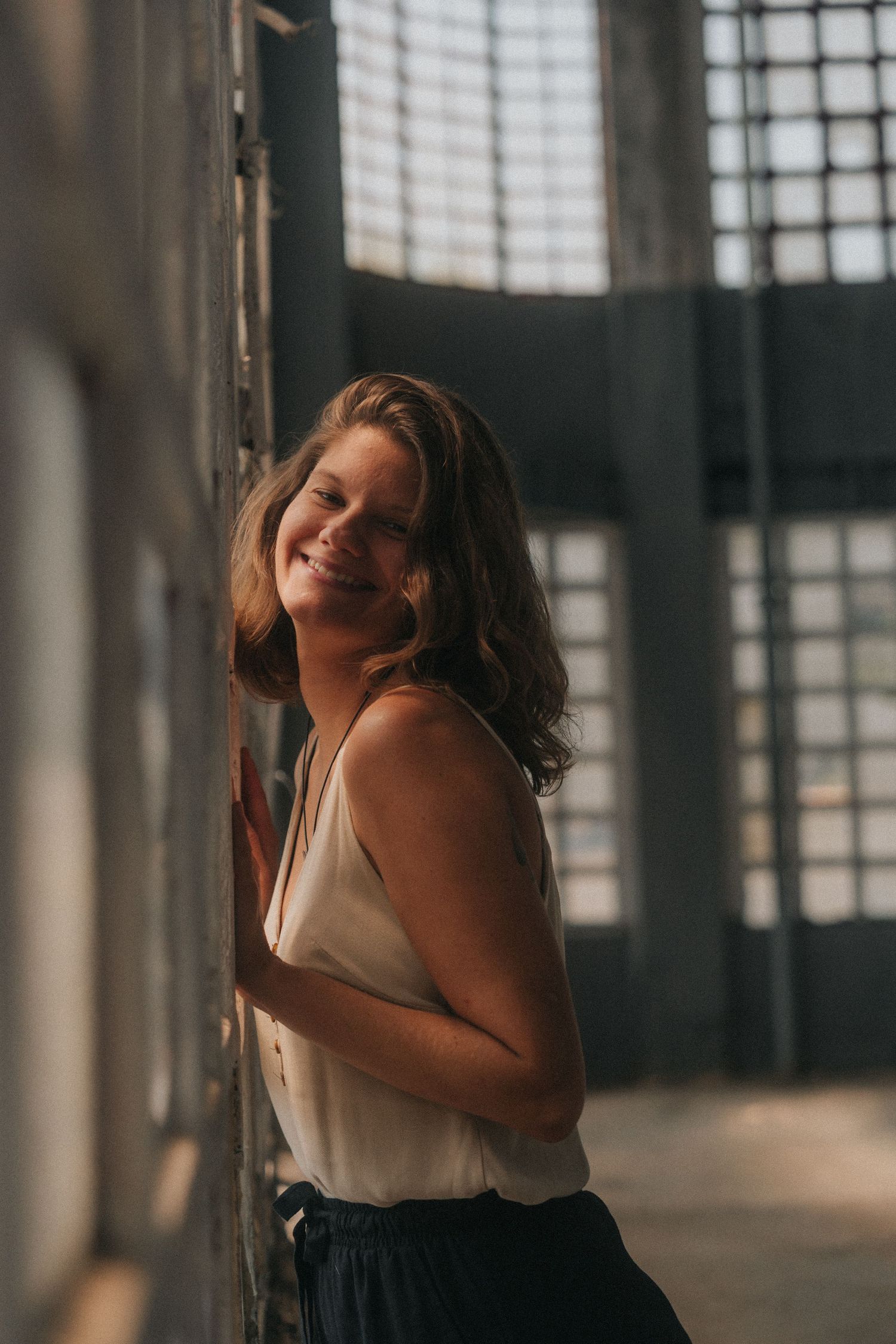Natural light portrait of someone in a white tank top and dark pants leaning against wall by window with moody lighting.