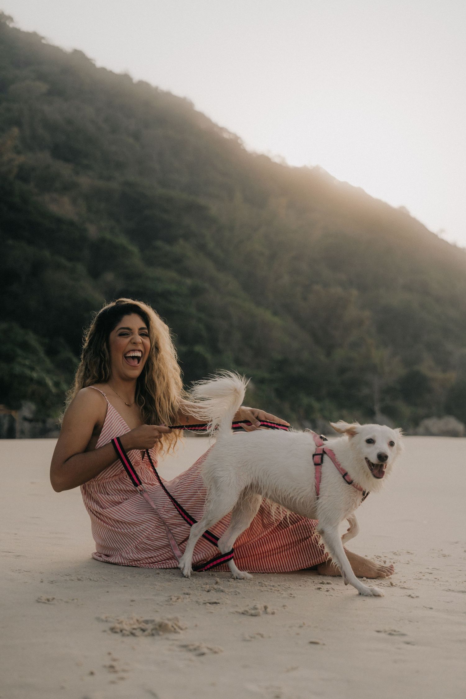 A woman in a pink dress playfully interacts with her white dog on a sandy beach at sunset.