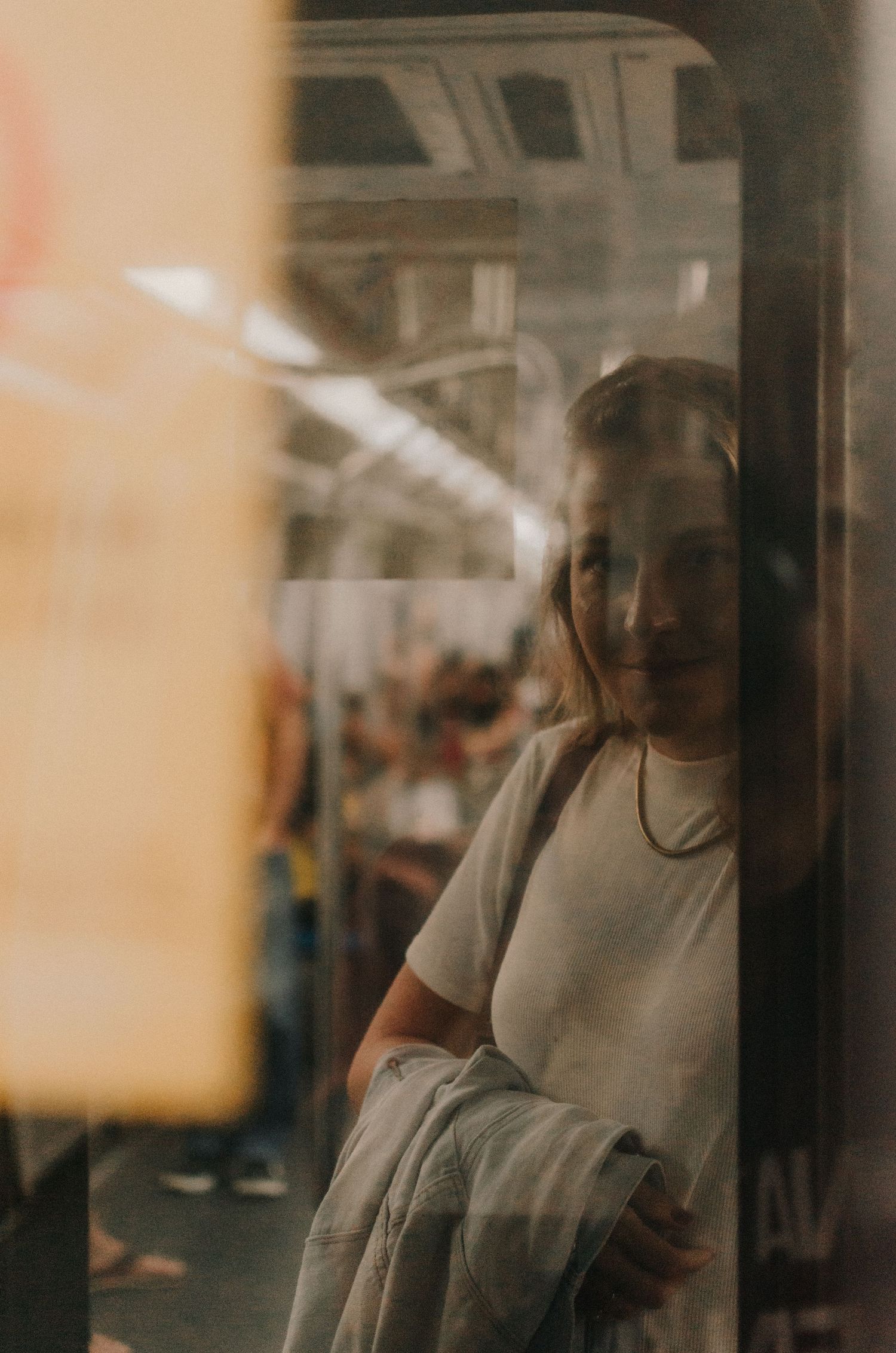 A person in a gray t-shirt is seen through glass reflections in what appears to be a subway or train environment.