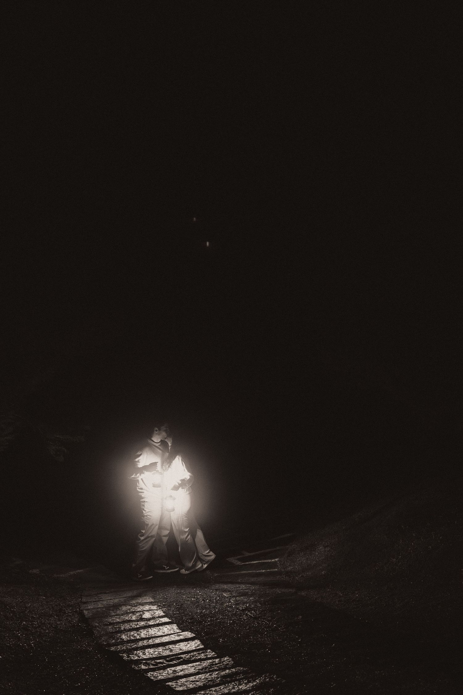 A glowing figure illuminates a dark wooden bridge or walkway at night creating an eerie atmospheric scene.