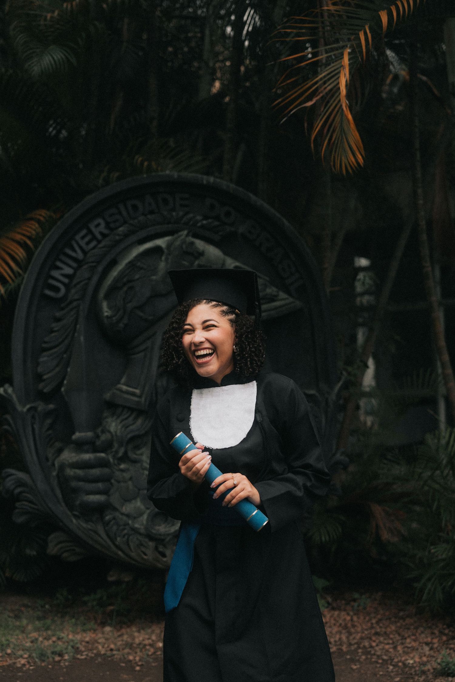 Graduate in traditional black cap and gown celebrates joyfully in front of a university emblem under palm fronds.