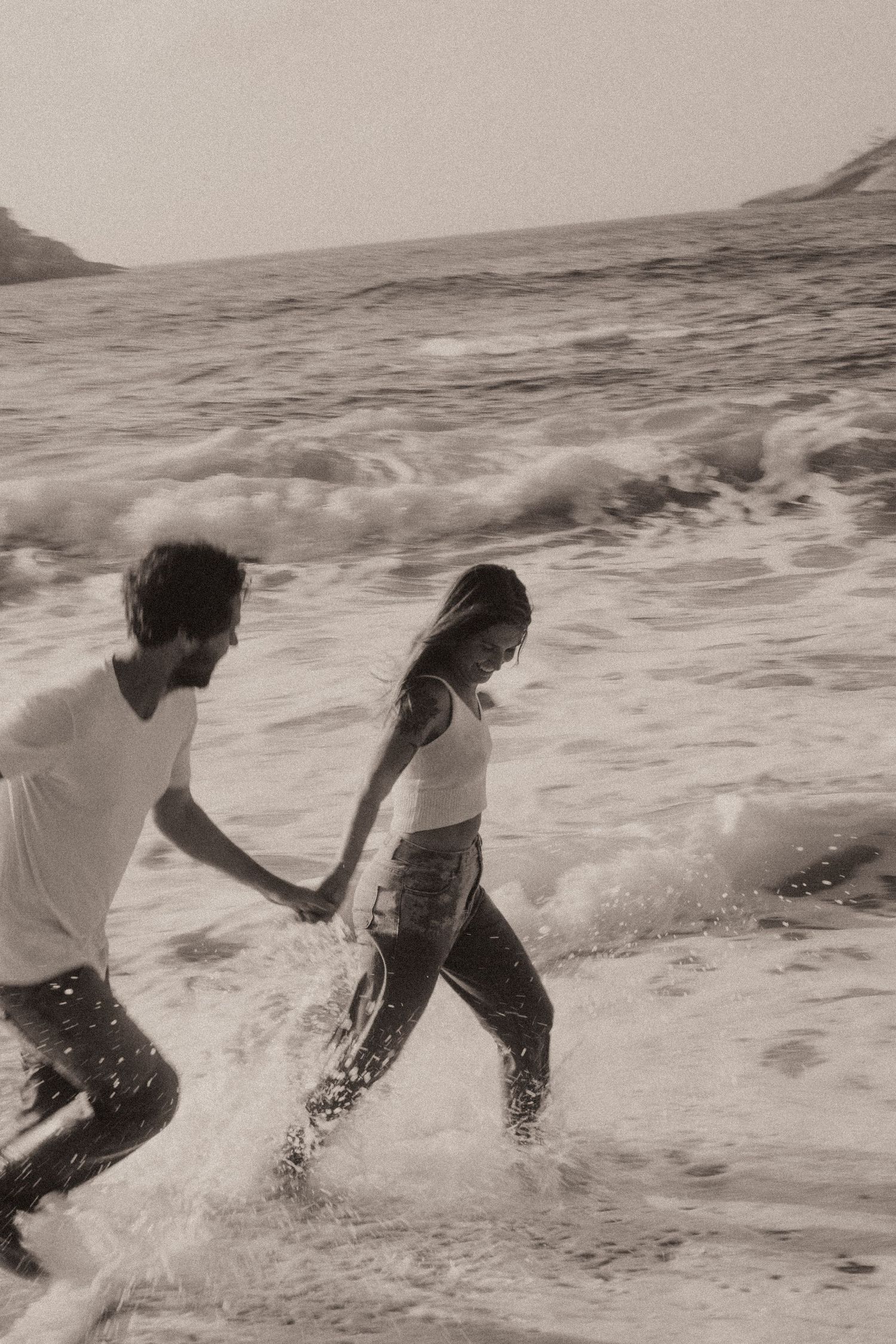 Two people playing together in the ocean waves as they break along the beach in black and white photography.