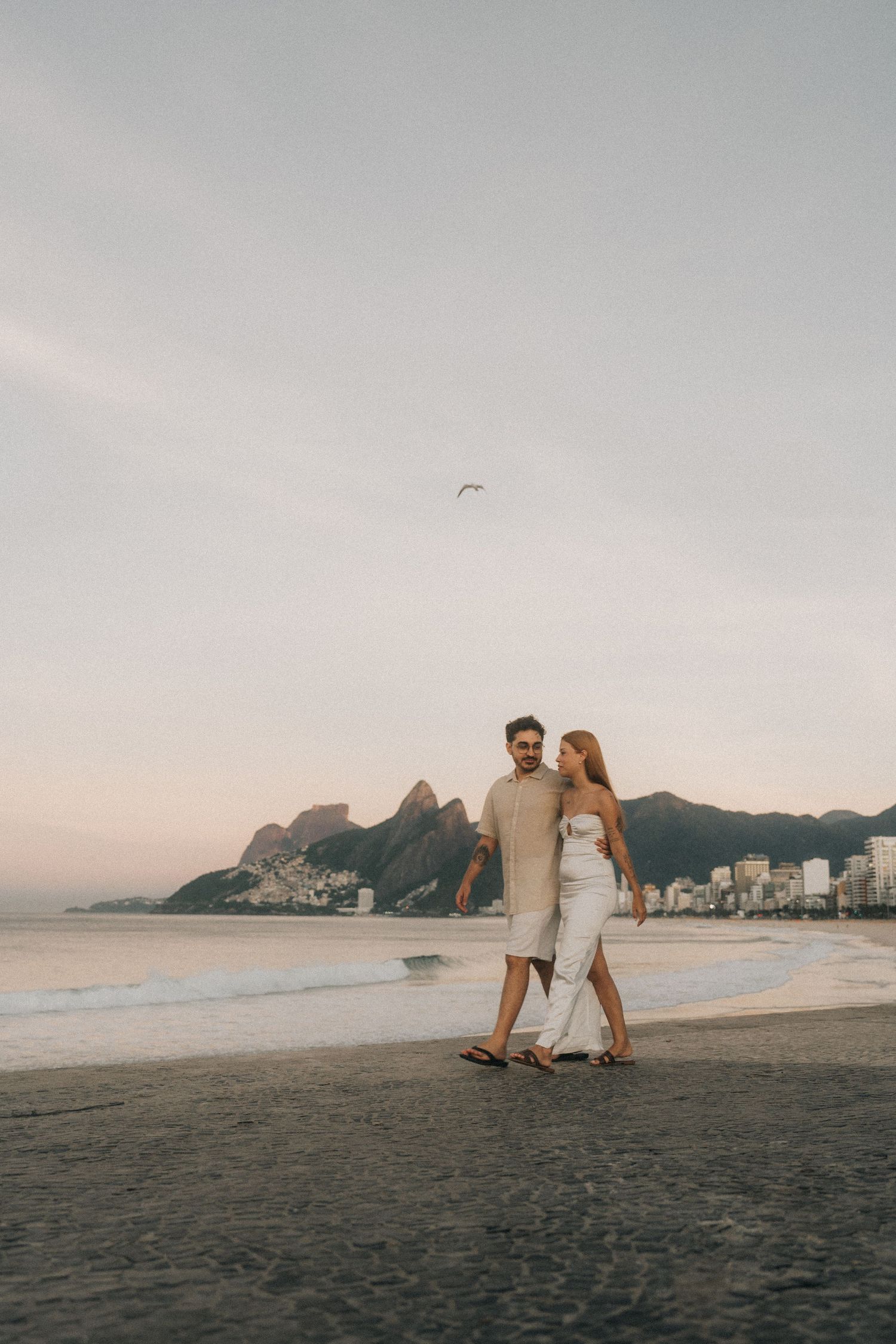 A couple walking together on Ipanema Beach in Rio de Janeiro with iconic Two Brothers mountains in the background at sunset.