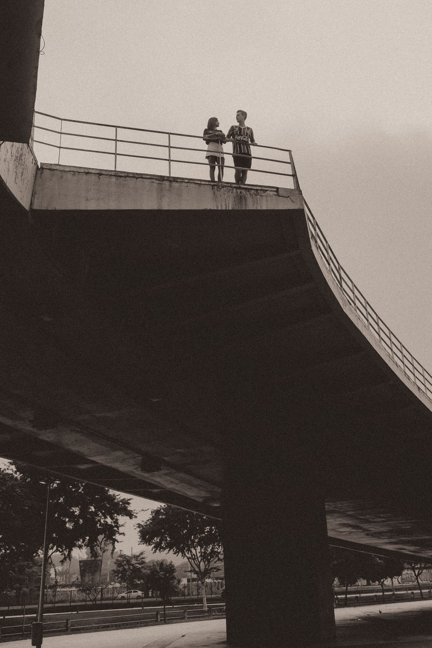 A black and white photograph shows figures observing from a bridge railing against an architectural backdrop.