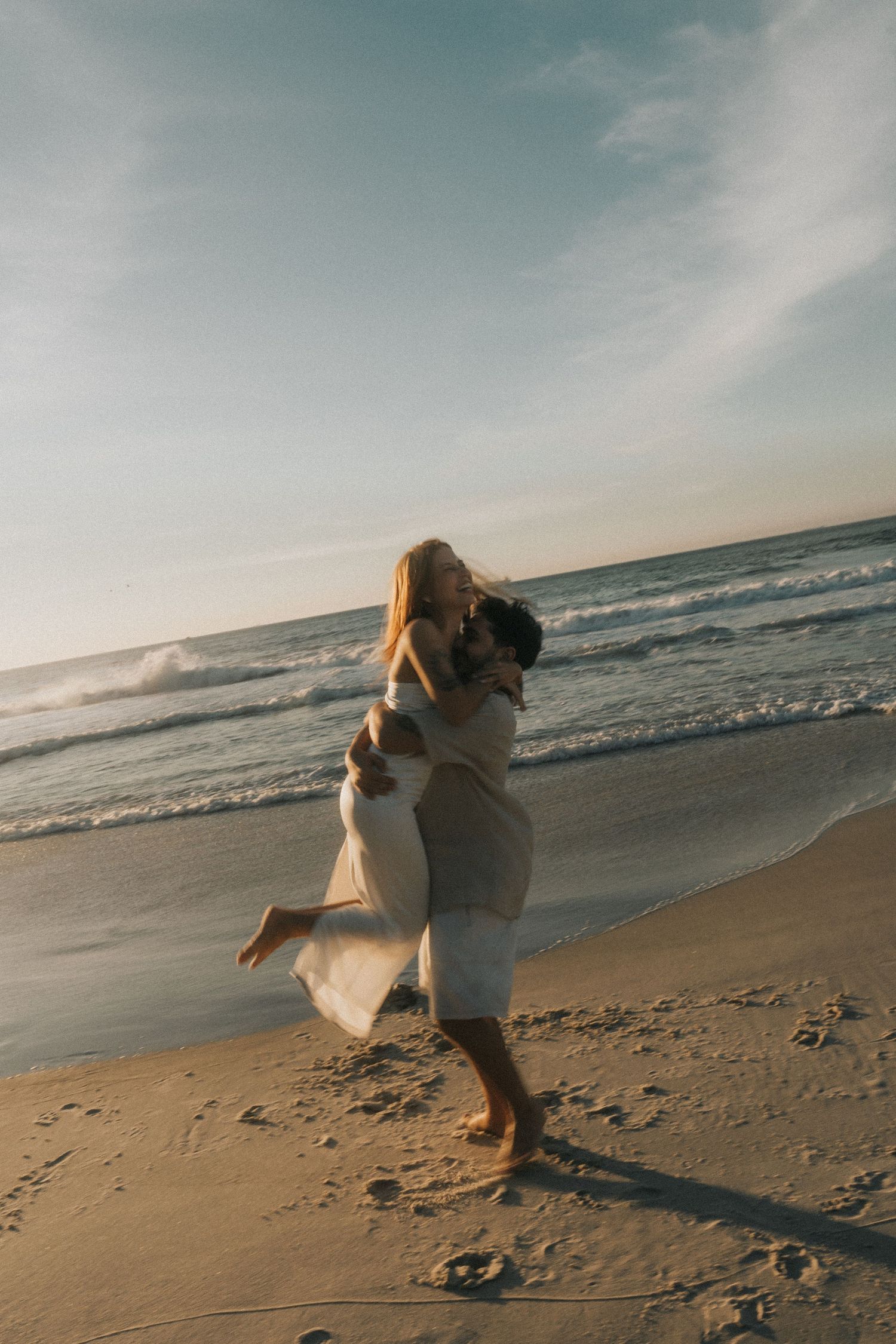 Two people dance together on a sandy beach at sunset with waves crashing in the background.