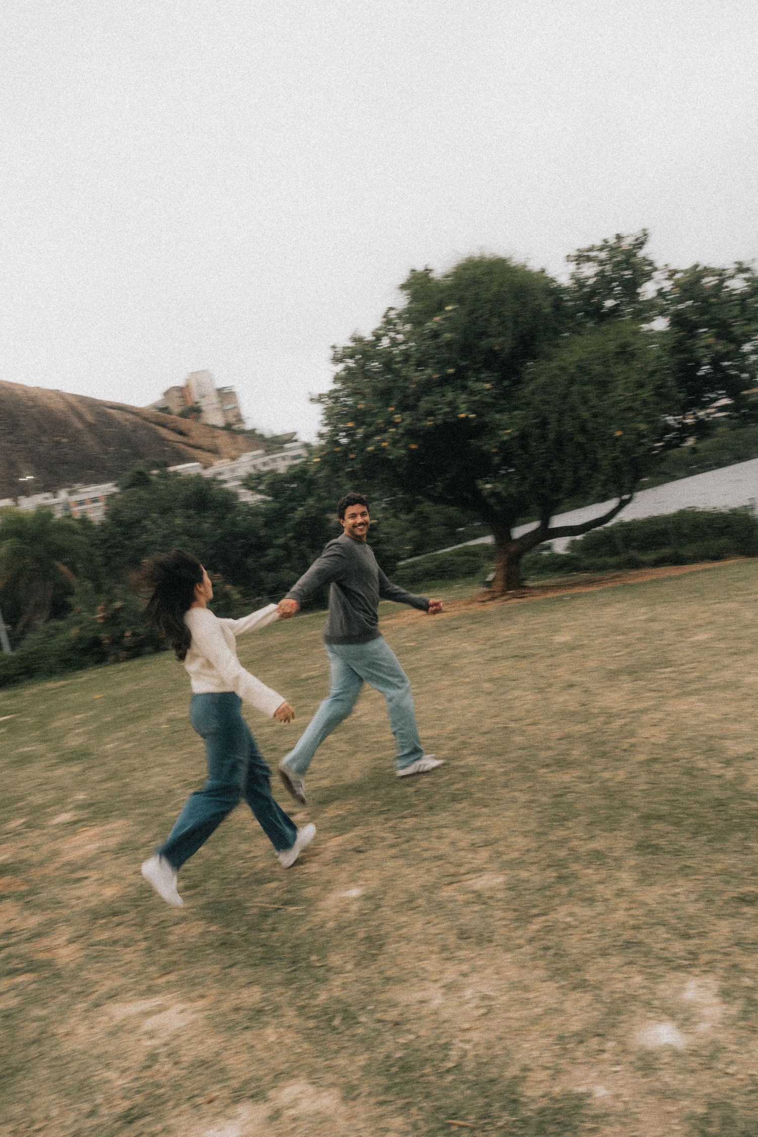 Two people playfully chasing each other in casual denim outfits across a grassy hillside on an overcast day.