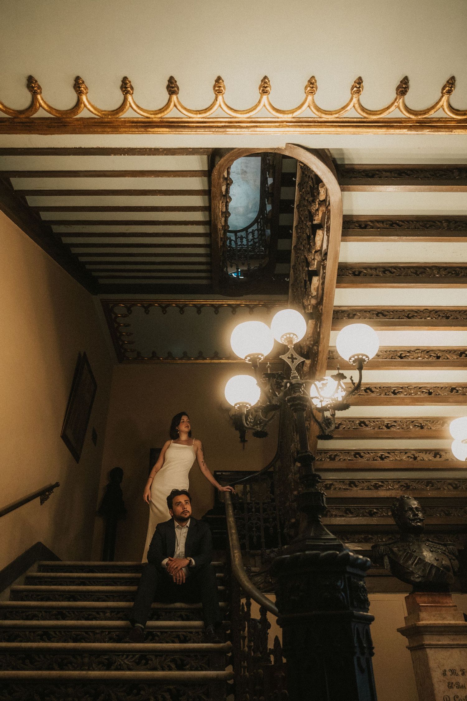 A couple poses on an ornate vintage staircase with decorative lighting and architectural details in a moody atmosphere.