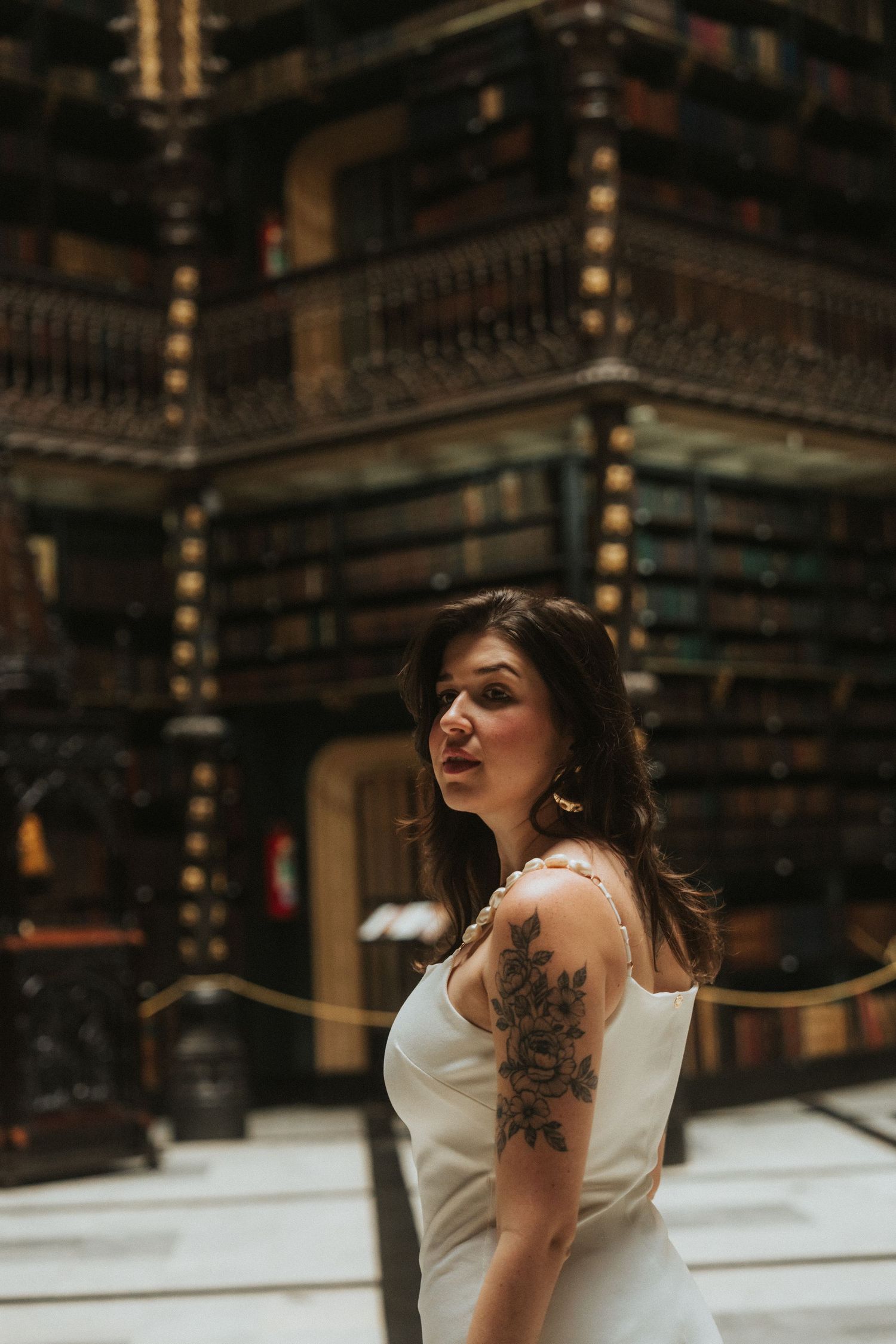 A bride in a white wedding dress poses dramatically in a grand library setting with ornate wooden bookshelves.