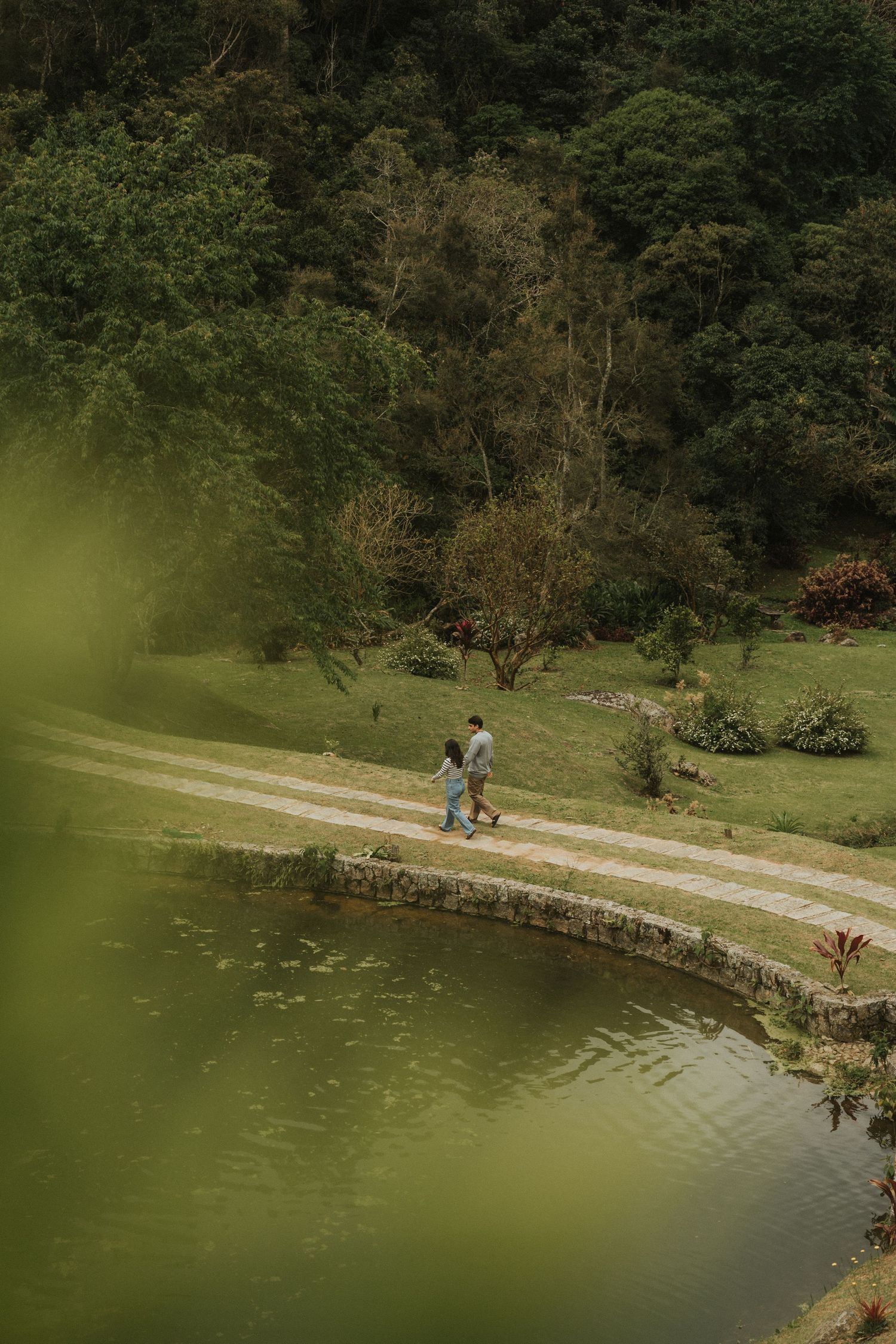 People walk along a curved stone wall path beside a serene lake surrounded by lush green forest on a summer day.