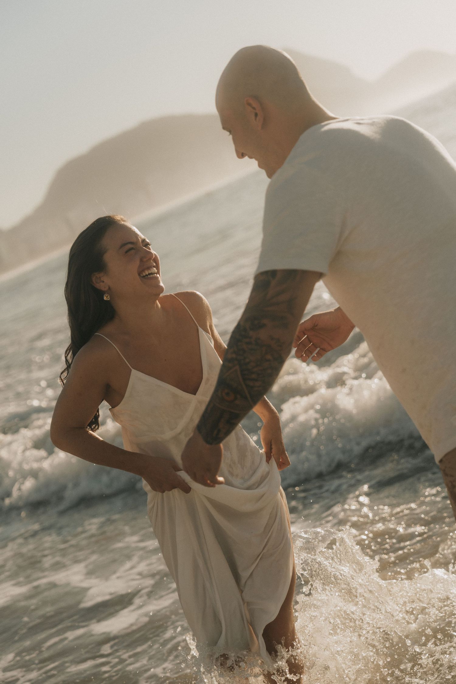 A joyful couple splashes in ocean waves at sunset, with the woman wearing a flowing white dress and sharing a candid moment.