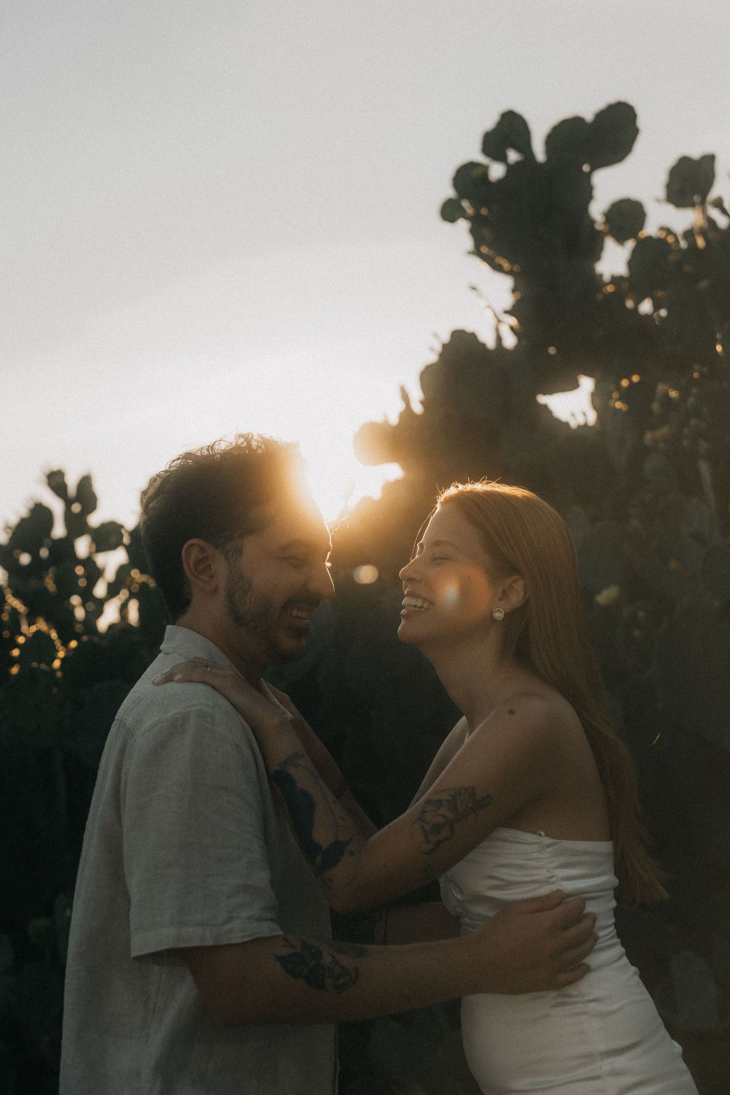 A romantic couple embraces at sunset in a garden setting with sunlight streaming through silhouetted trees.