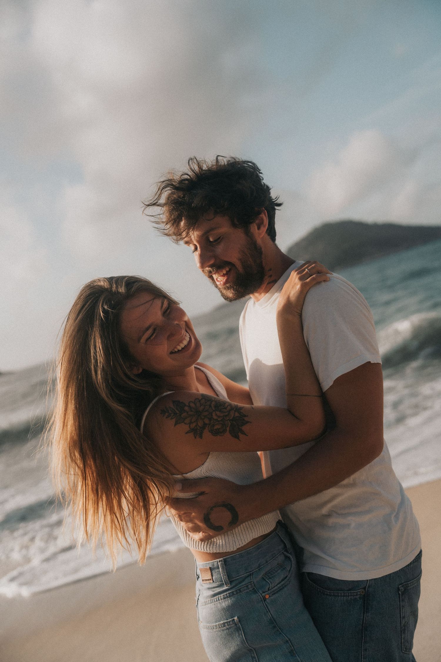 A joyful couple in casual outfits laughs and embraces together on a sunny beach with waves in the background.