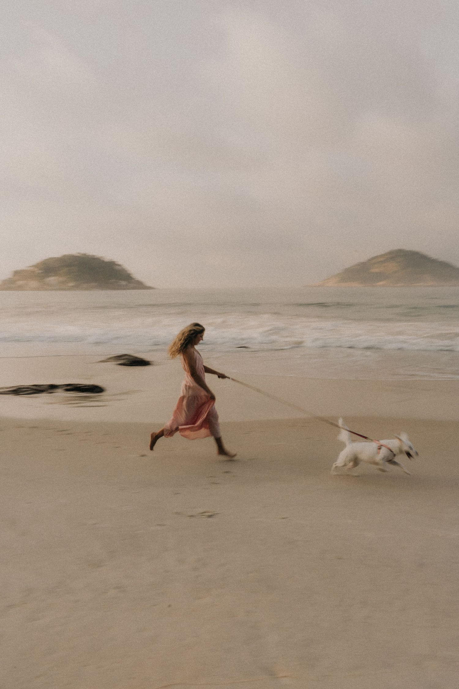 A person runs along a misty beach with a white dog against a backdrop of distant islands and overcast sky.