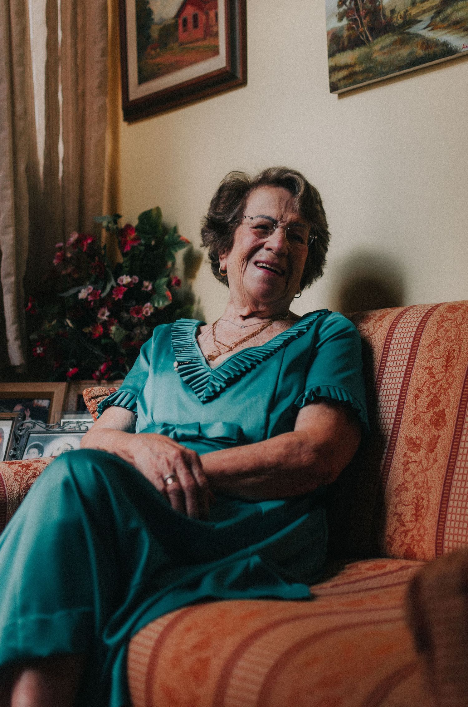 A person wearing a turquoise outfit relaxes on an orange floral patterned couch in a living room with framed artwork.